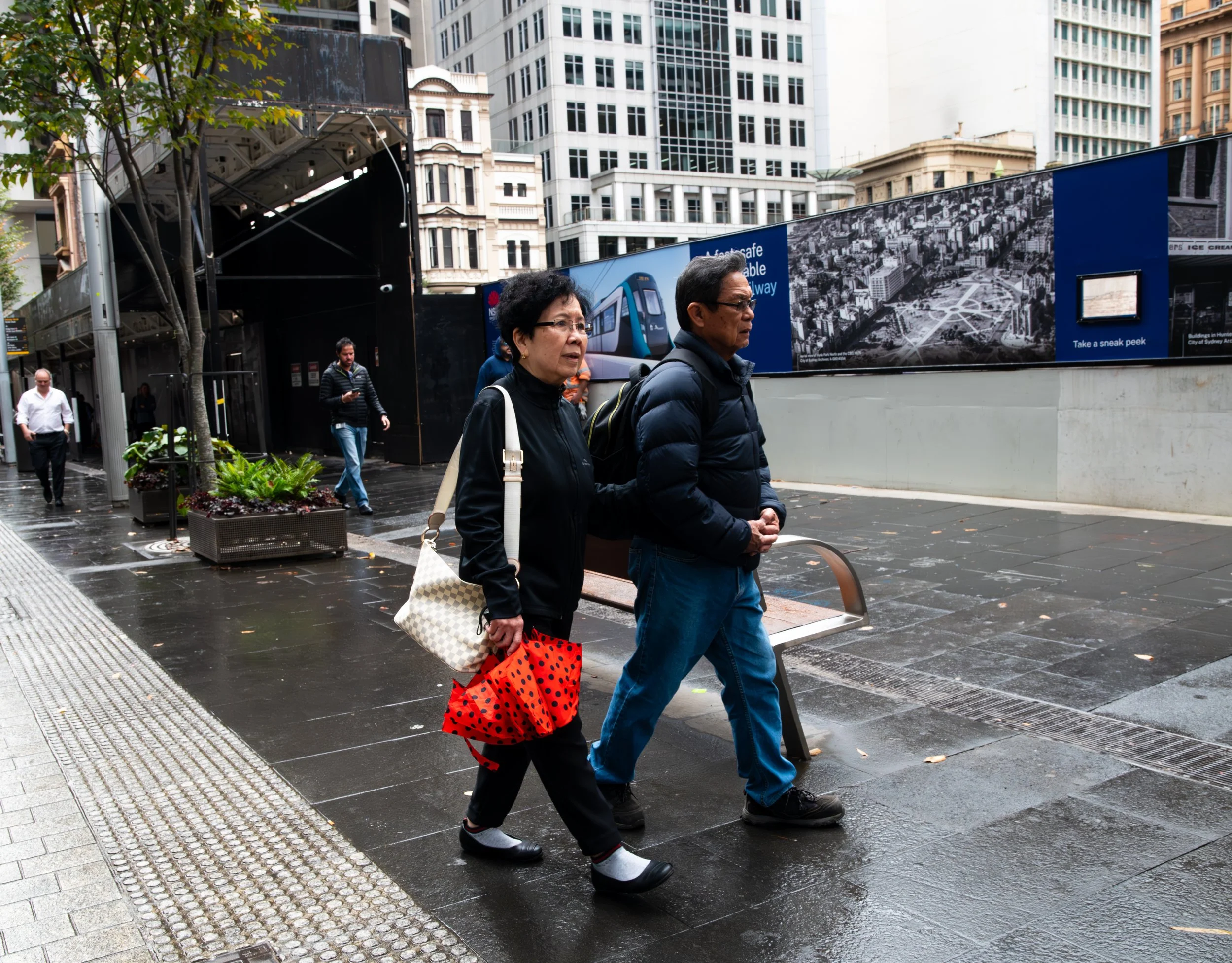 A man and woman walking on a wet city sidewalk with tall buildings and advertisements in the background.