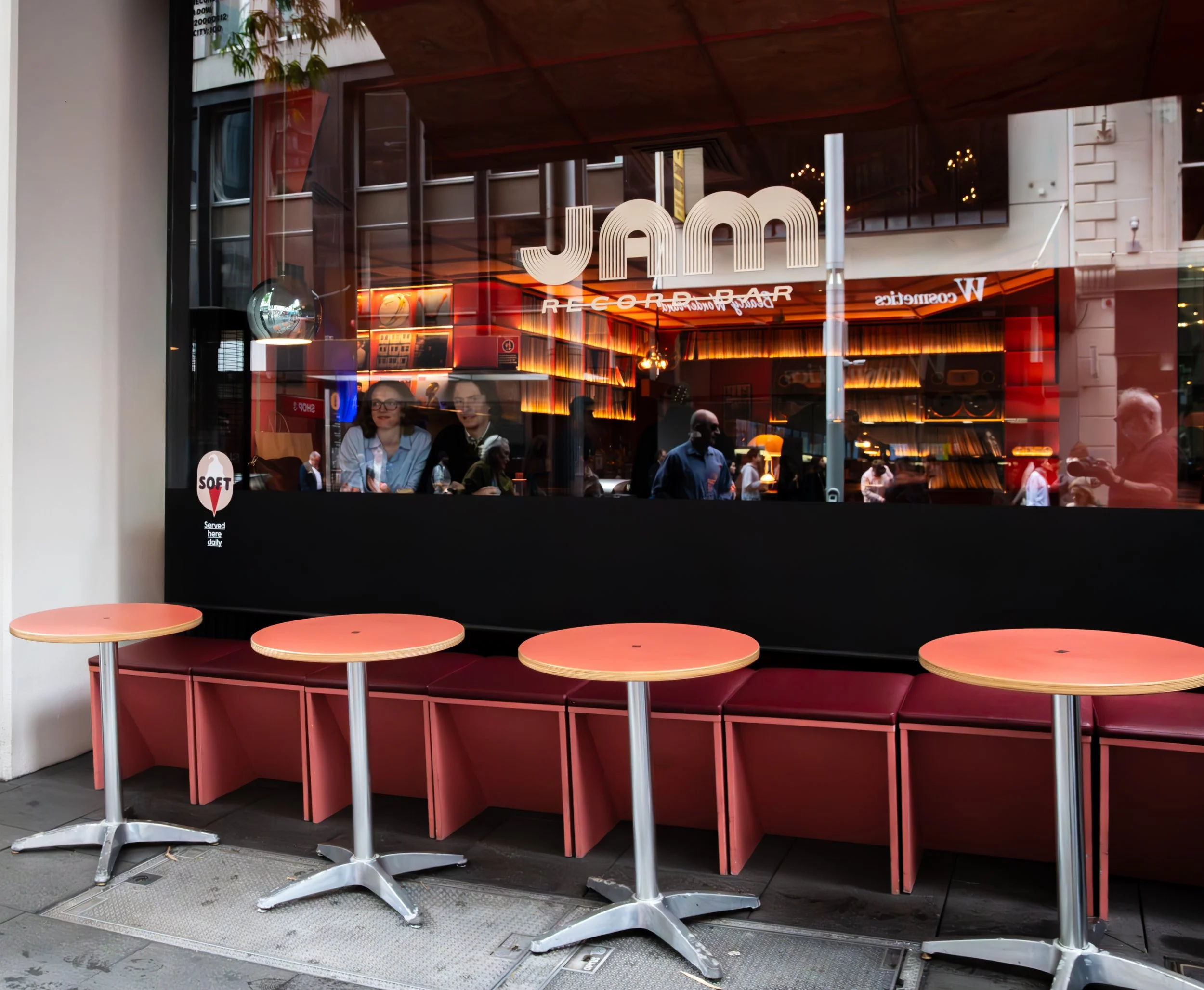 Empty pink tables with red bench seating outside a cafe, reflected in a glass window that shows people inside and a neon sign reading "JAMM" in the background.