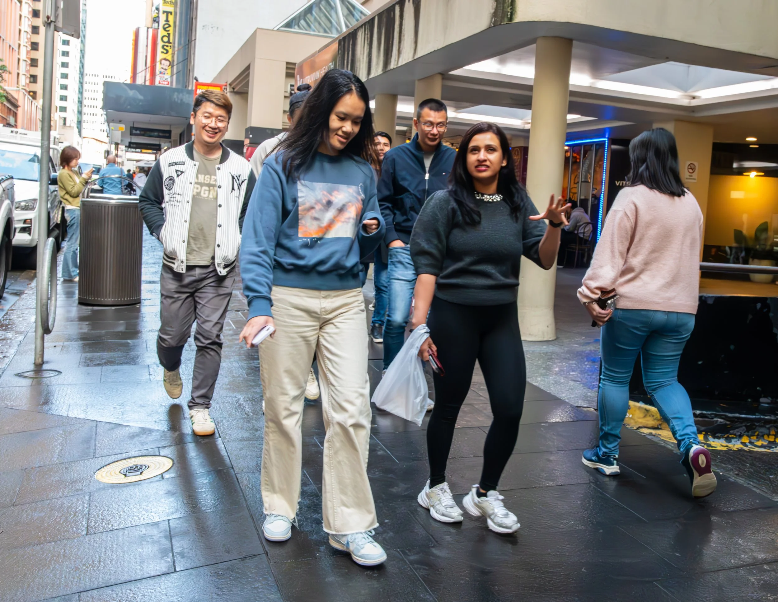 A group of young adults walking on a city sidewalk, talking and smiling, in an urban environment with taller buildings, shops, and street signs in the background.