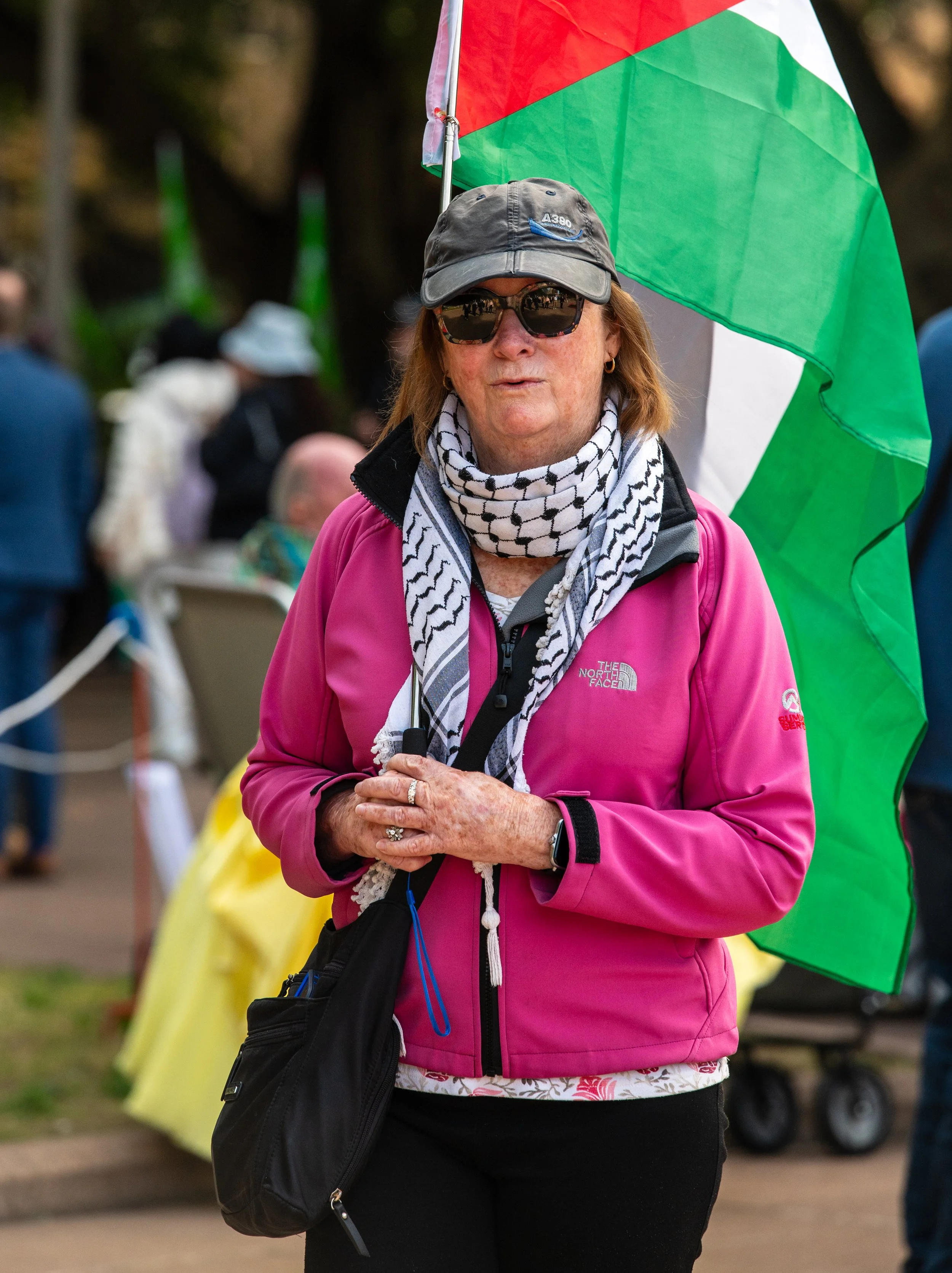 A woman wearing sunglasses, a gray cap, a pink jacket, and a black-and-white checkered scarf stands outdoors at a public event, holding a Palestinian flag.