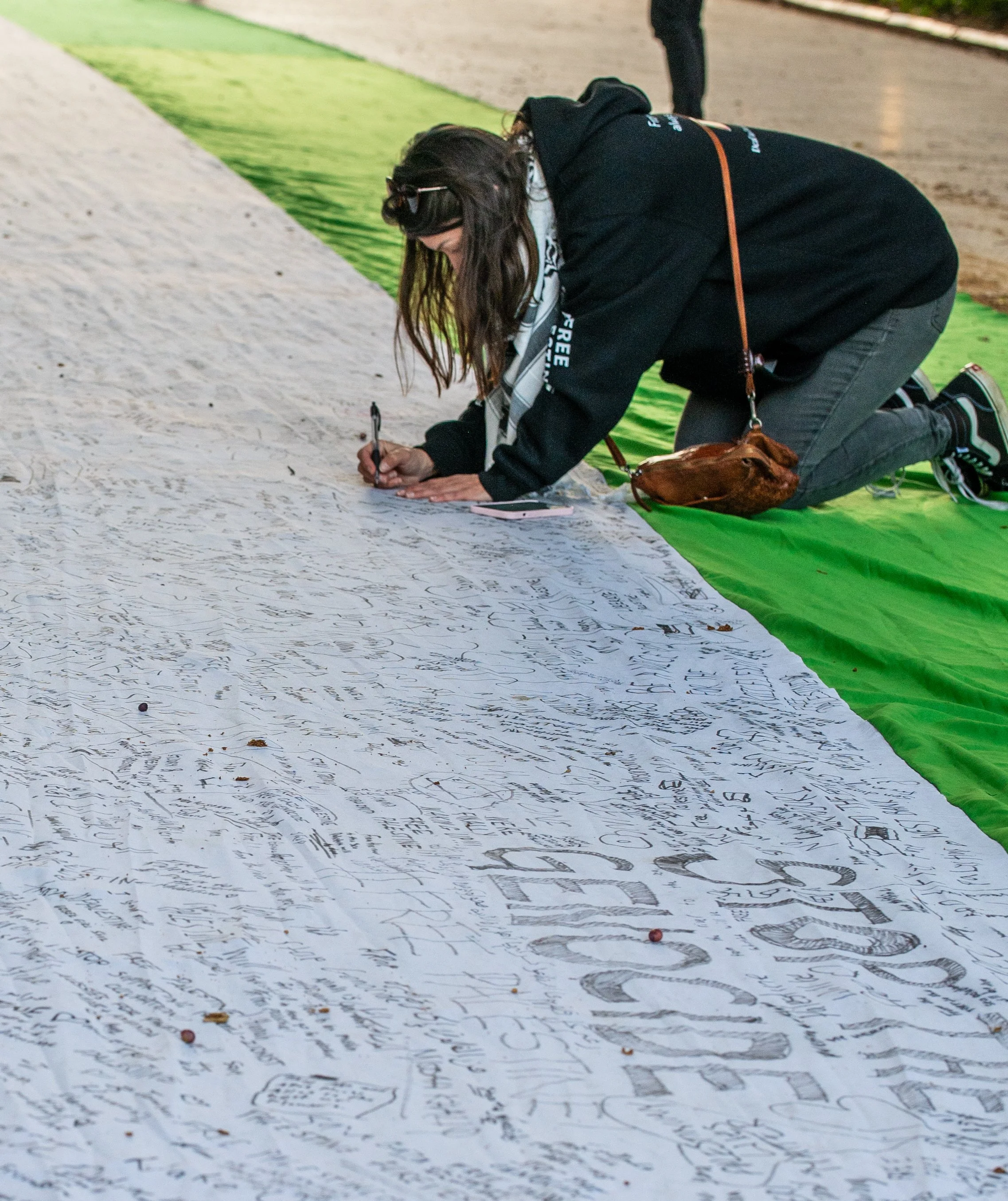 A woman in black hoodie and gray pants kneels on green mat, writing on a long white banner with black markers, at an outdoor event.