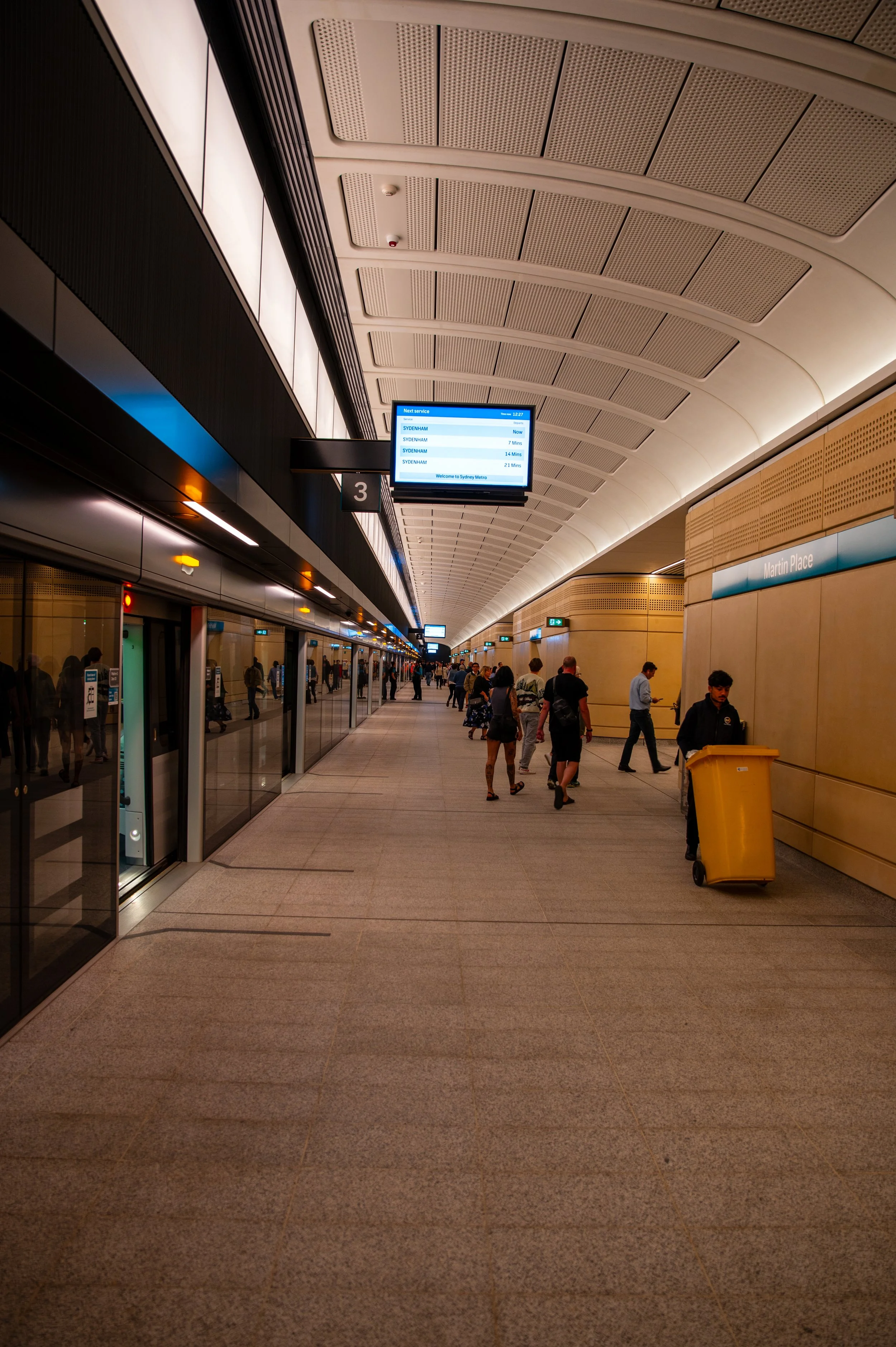 People walking on a tunnel-like train station platform with digital departure boards overhead and a sign indicating platform 3. A person with a yellow trash bin is visible on the right.