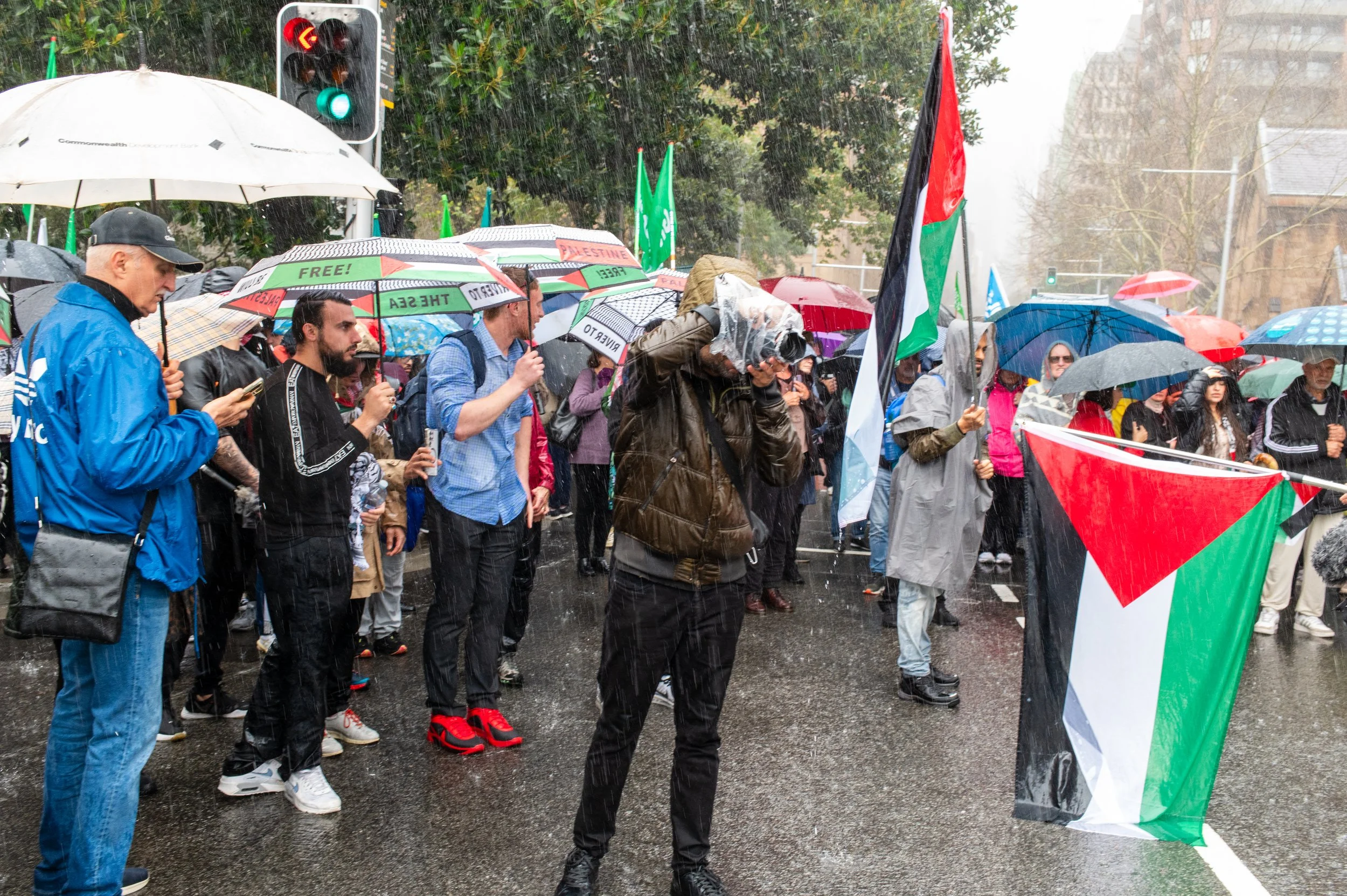 Protestors holding Palestinian flags during a rainy day protest, many with umbrellas, on a city street.