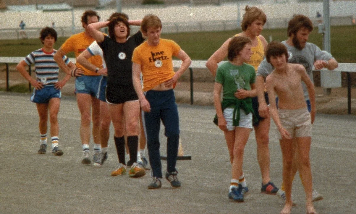 Group of children and teenagers at an outdoor track, some looking tired or bored, dressed in athletic clothing from the 1970s.
