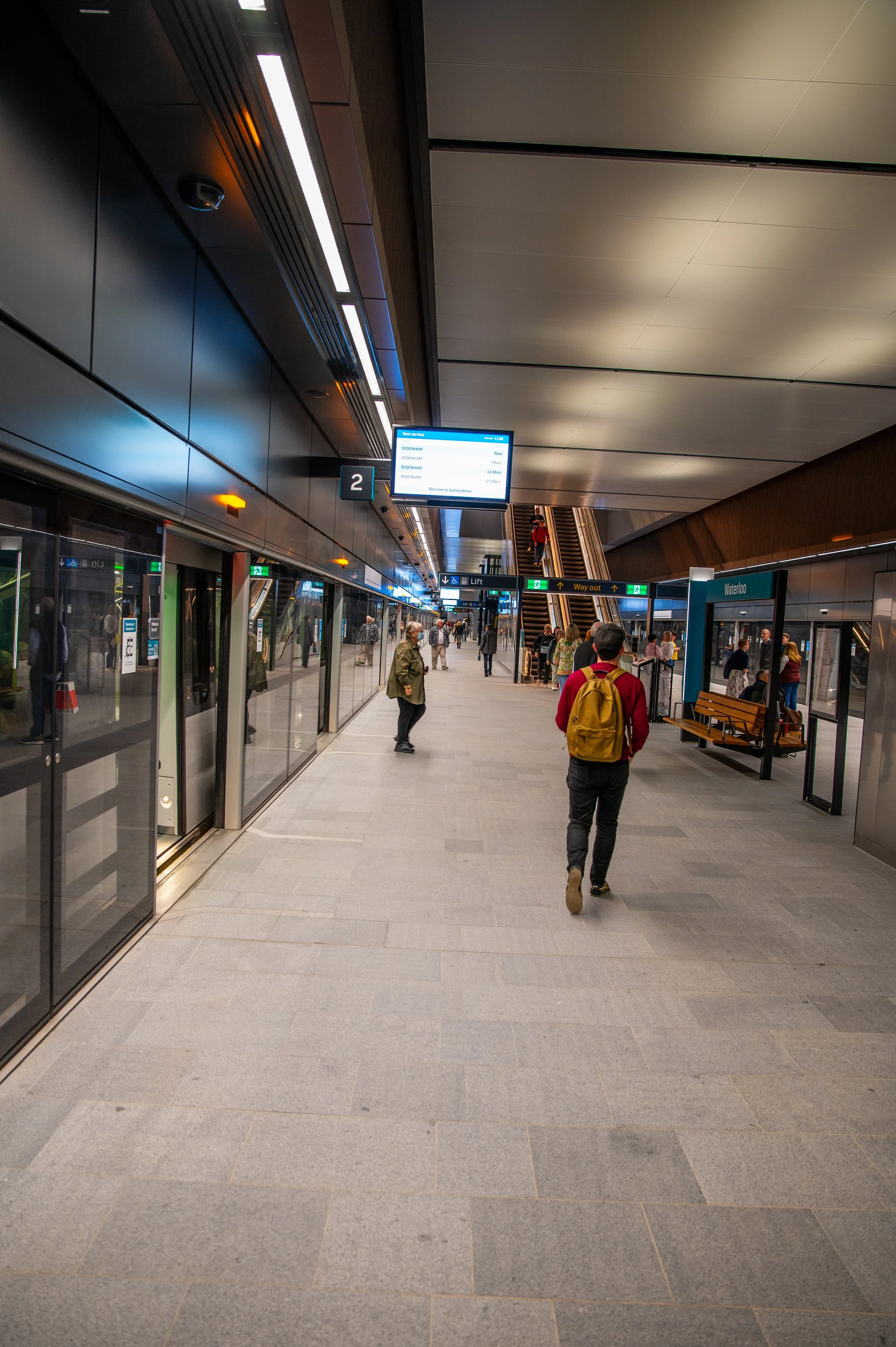 People waiting on a train platform with an electronic departure board overhead, a staircase, and a waiting area with benches.