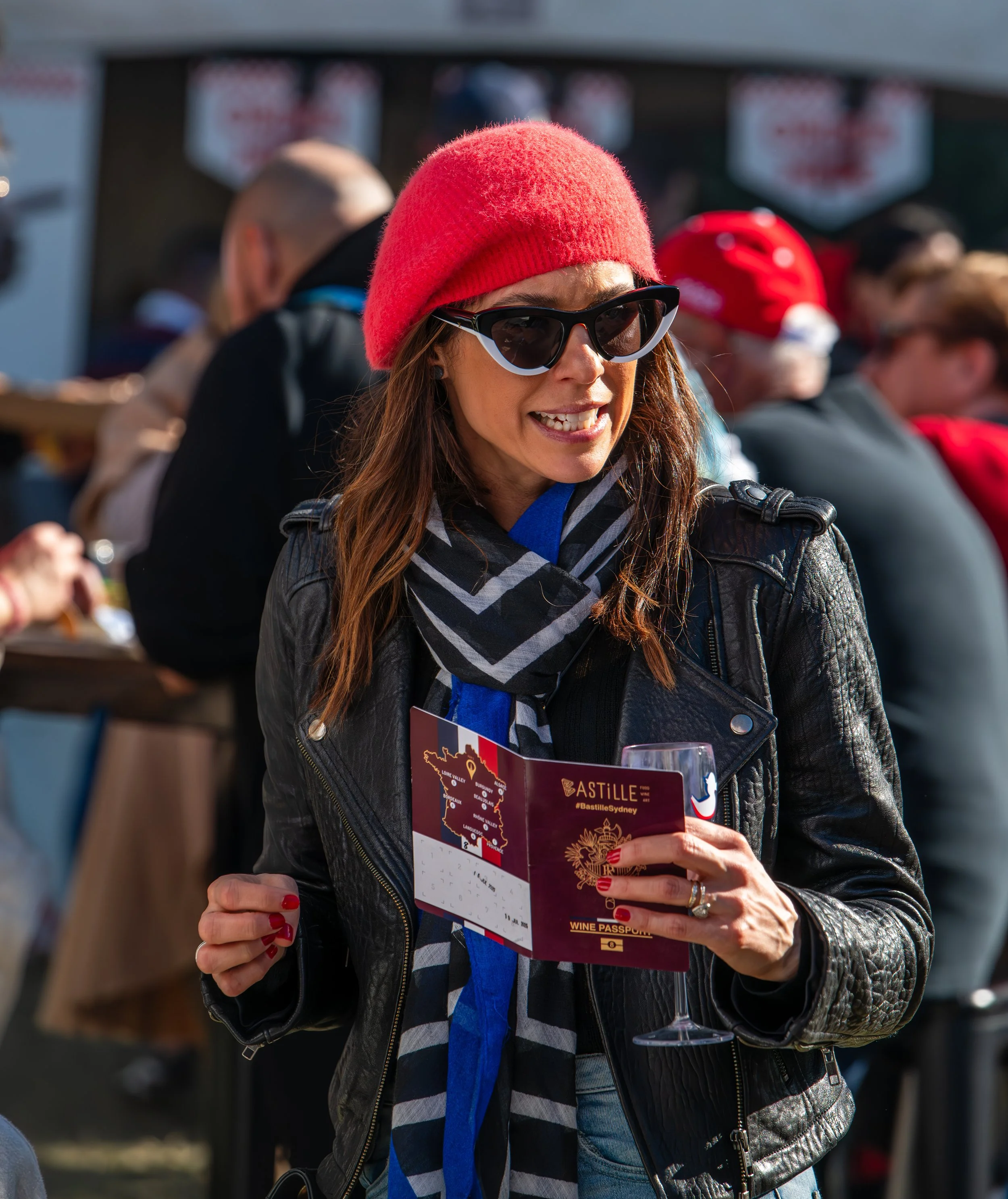 A woman wearing a red beanie, black jacket, and sunglasses holding a wine passport at an outdoor event with a crowd in the background.