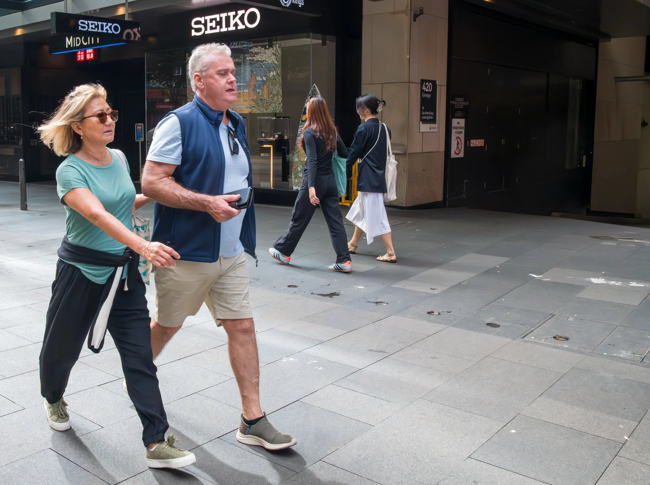 A group of four people walking on a city sidewalk in front of store fronts, including a woman with sunglasses and a man in a blue vest holding a phone. Two women are walking in the background, one with a white skirt and the other with black pants, both carrying bags.