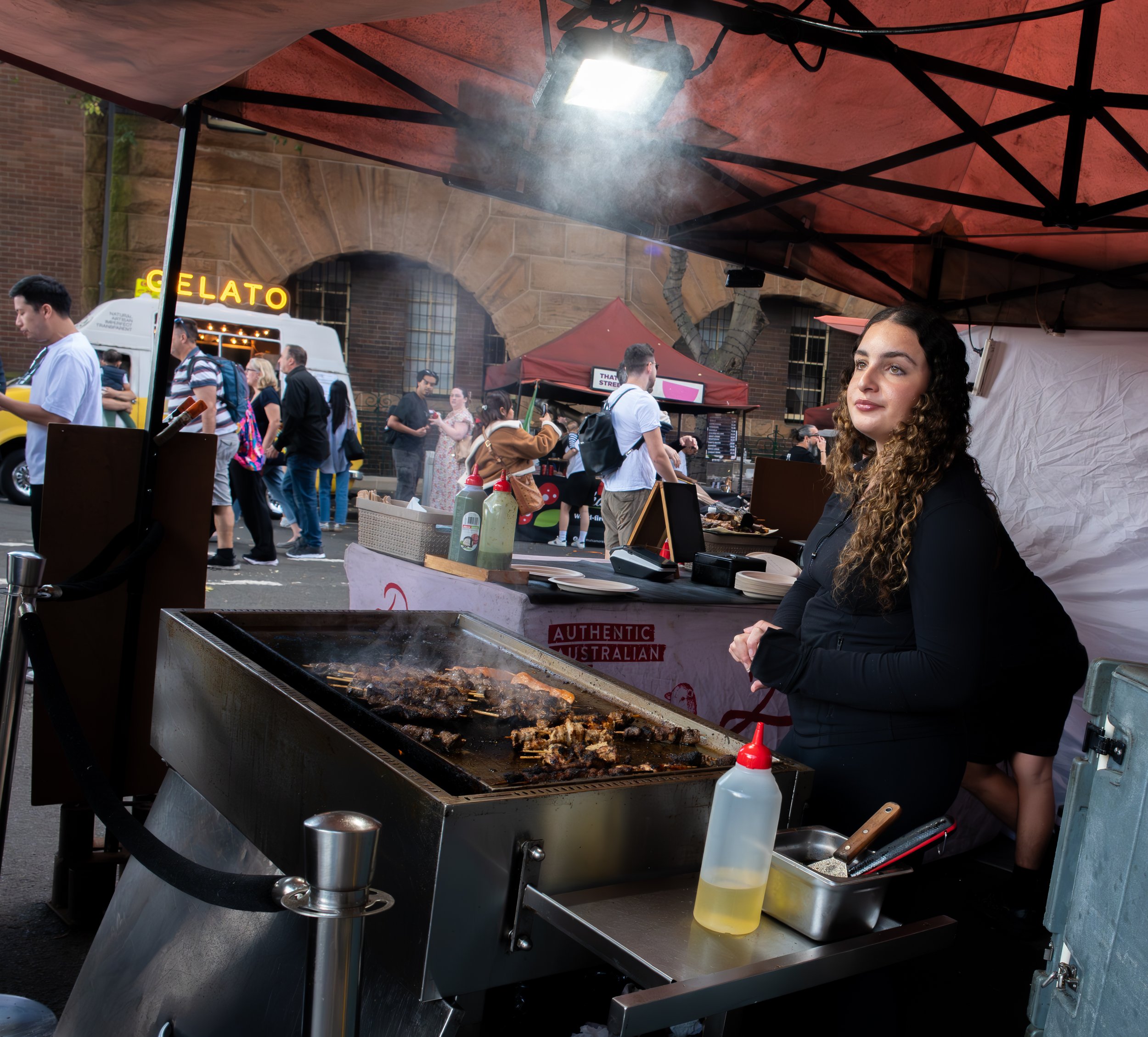 A woman standing behind a grill at a food stall, with a busy street scene in the background including a gelato truck, pedestrians, and market stalls, under a red canopy.