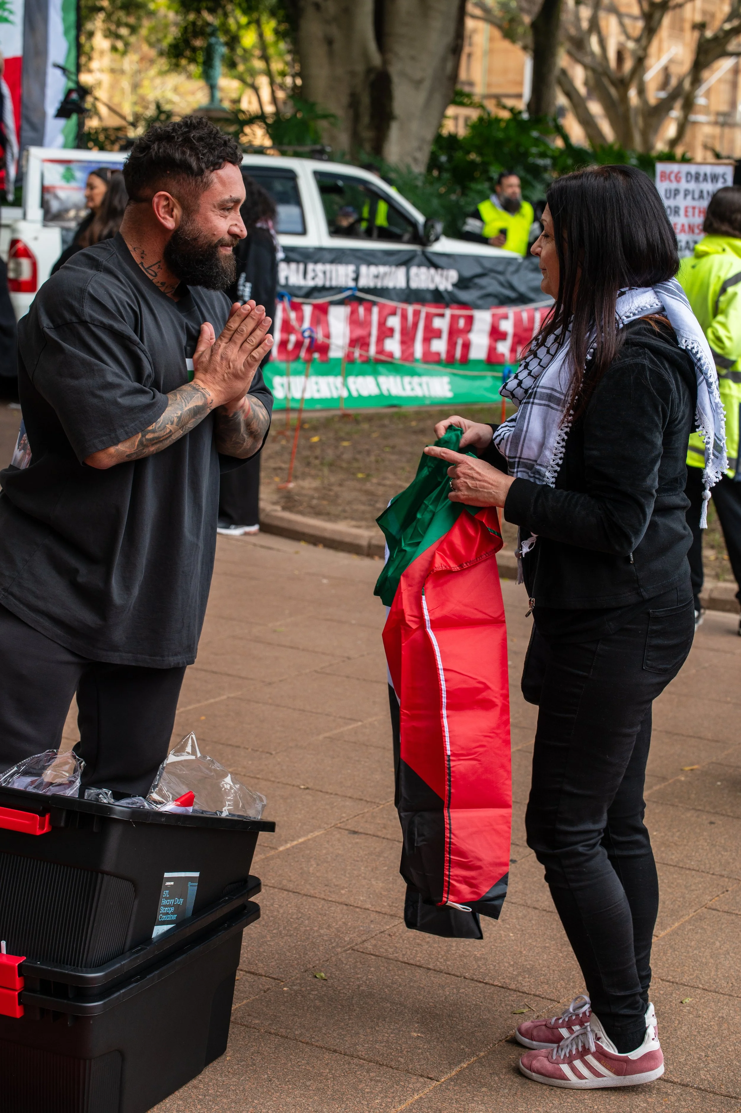 A man with a beard and tattoos on his arm is smiling and holding his hands together in front of a woman at an outdoor protest. The woman, wearing a black jacket and a scarf, is holding a folded Palestinian flag. In the background, there is a truck with a banner that reads 'Palestine Action Group' and 'Never End' and other people wearing yellow vests.