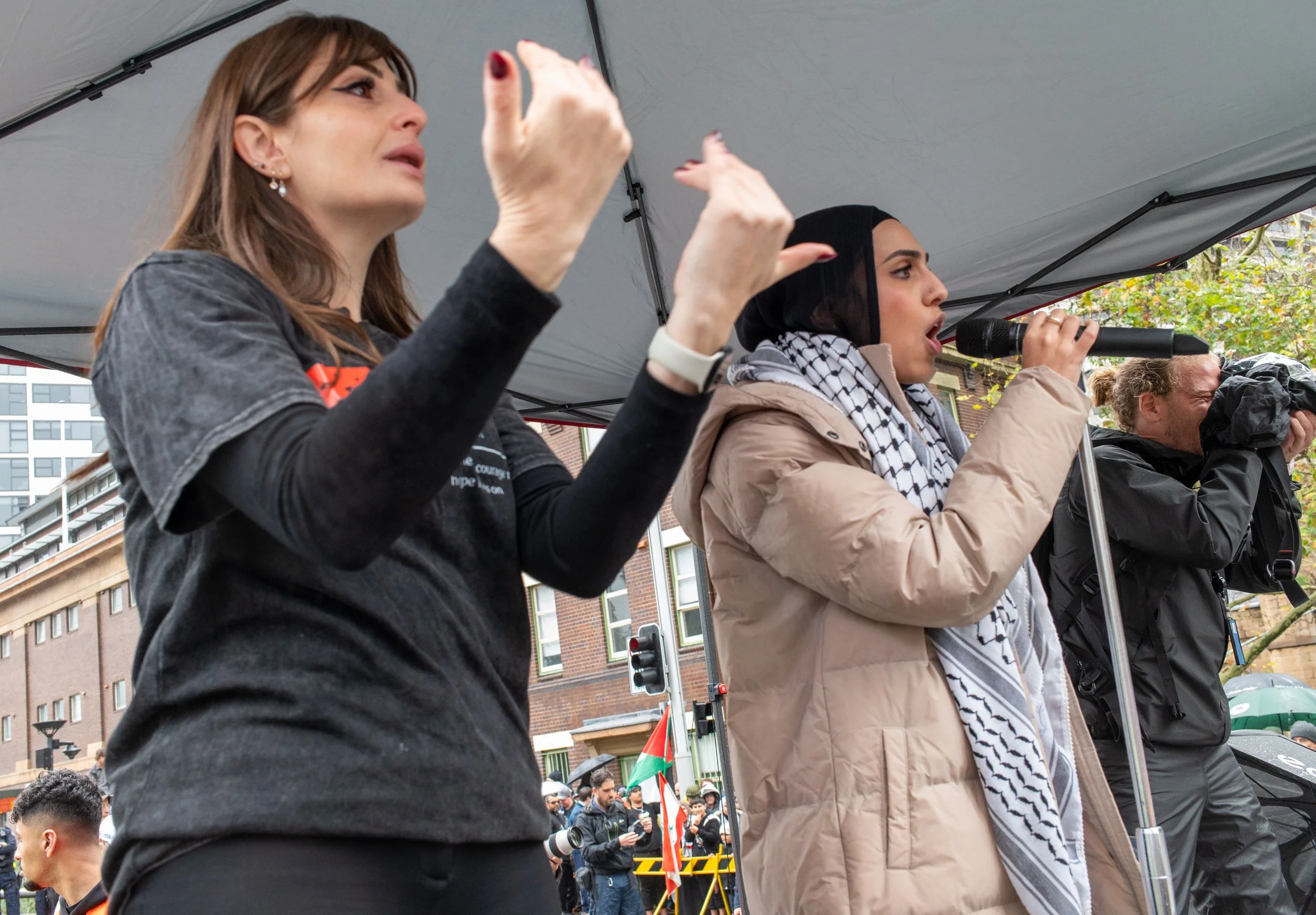 Three women protesting outdoors under a tent, with buildings and a crowd in the background. The woman on the left has light skin, brown hair, and is wearing a black t-shirt, raising her hand. The woman in the middle has light skin, dark hair with a headscarf, and is speaking into a microphone. The woman on the right has light skin, blonde hair, and is holding a camera.