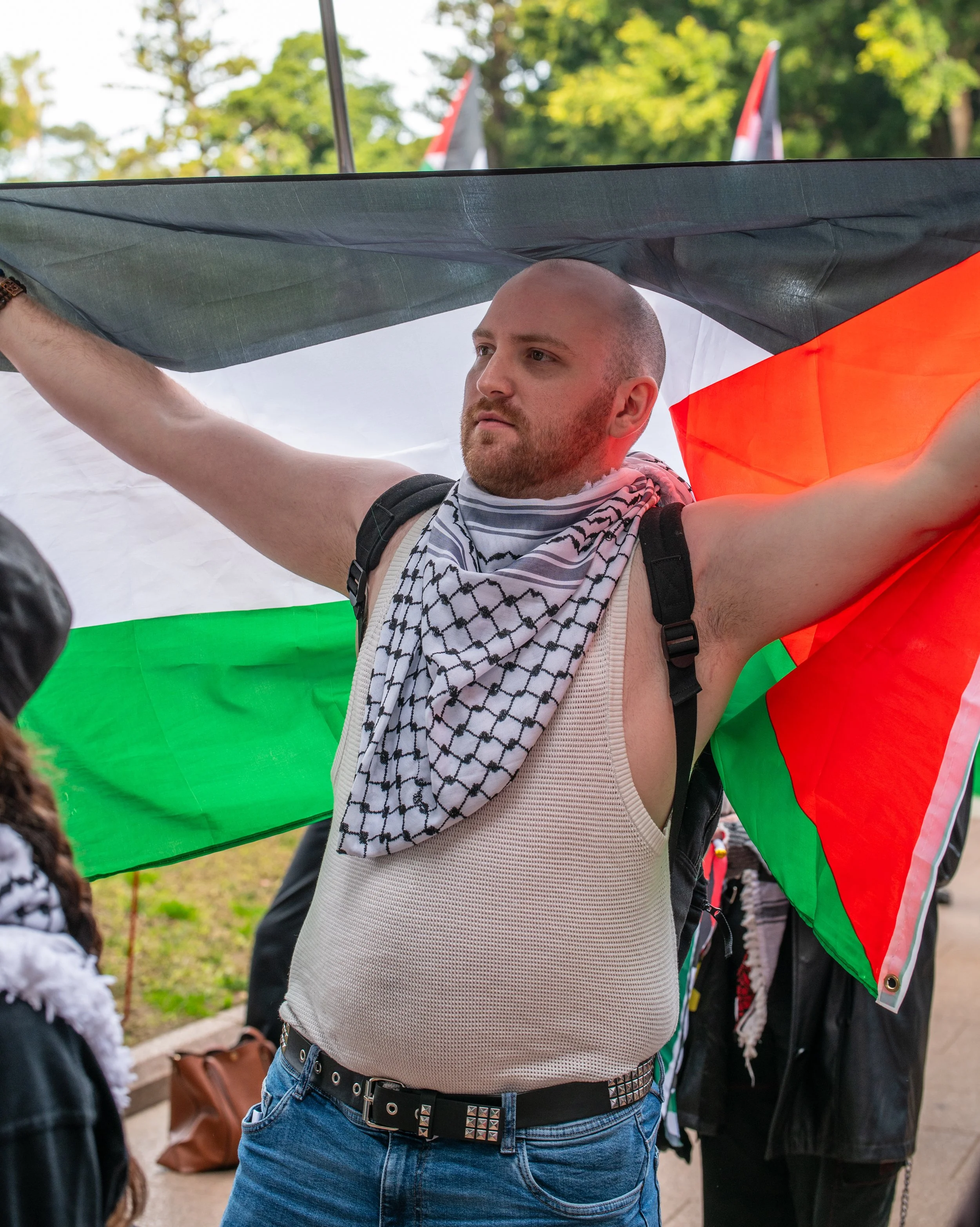 A man with a shaved head and beard, wearing a sleeveless shirt, is holding a Palestinian flag draped across his shoulders at a rally or protest.