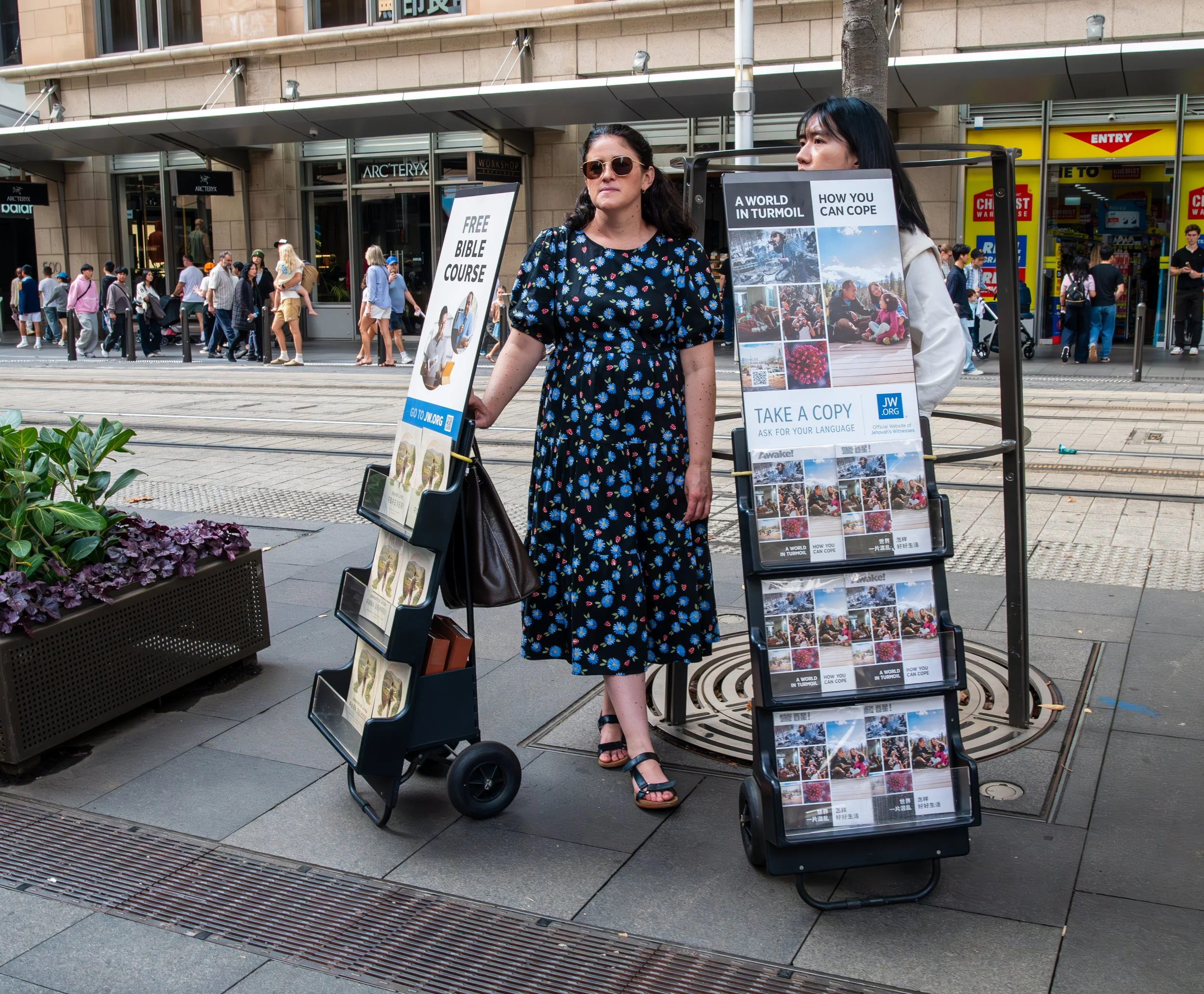 A woman standing on a city sidewalk next to two signboards promoting a free Bible course, with a busy street and shopfronts in the background.