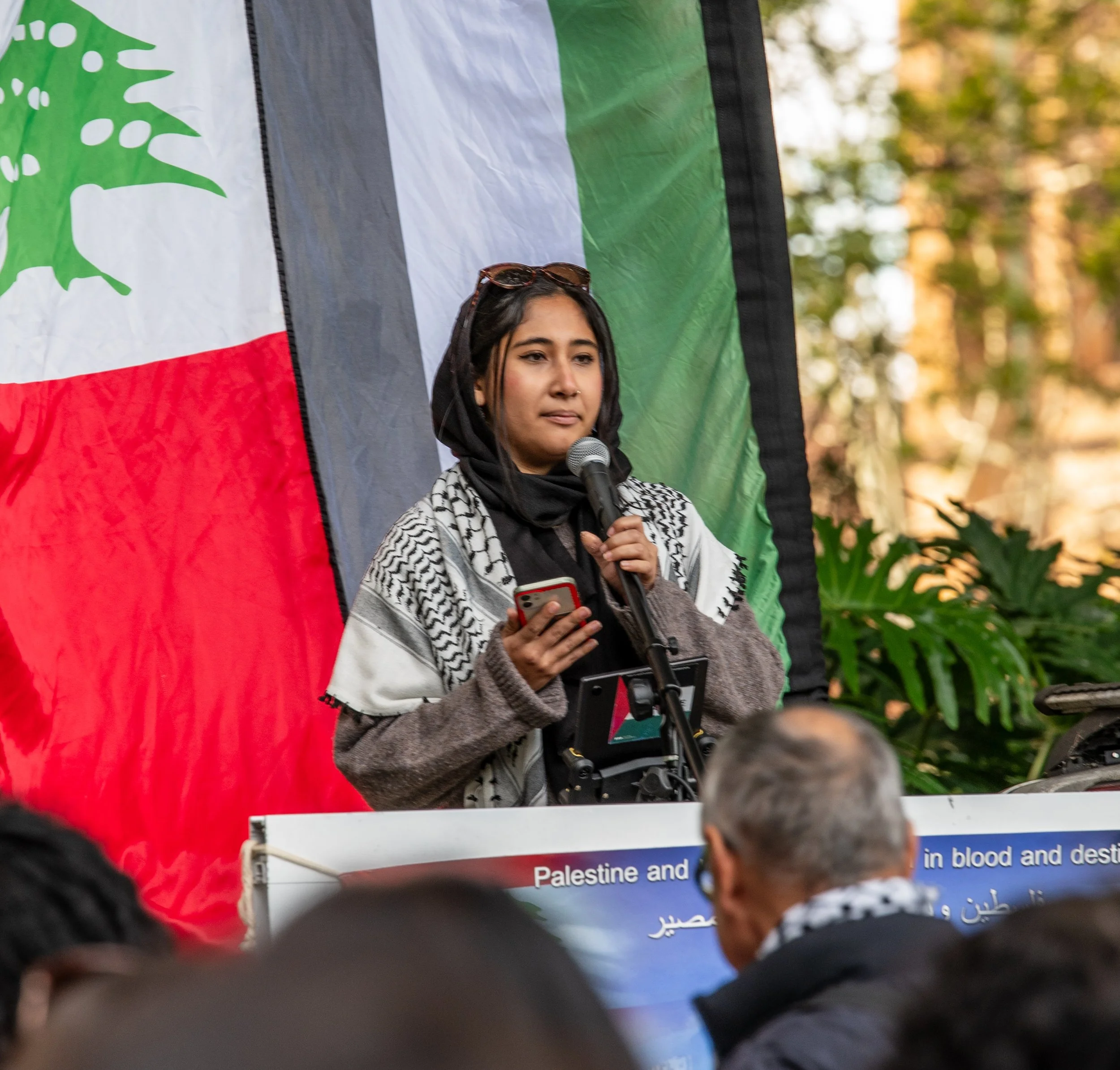 A woman speaking into a microphone at a rally or protest with a Palestinian flag behind her, holding a smartphone in her hand.
