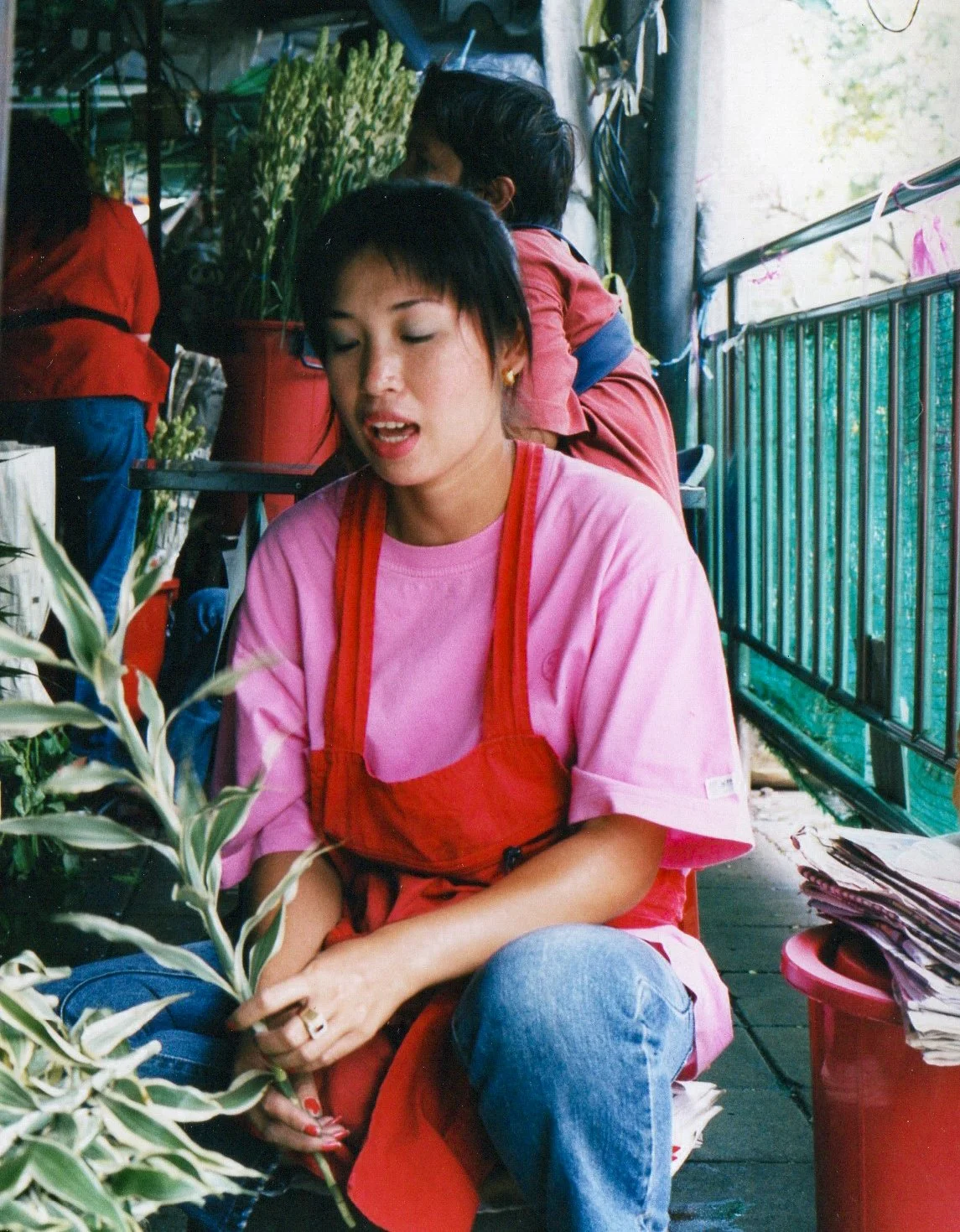 A young woman with short black hair wearing a pink shirt and red apron sitting on a chair at a plant shop, surrounded by plants and garden supplies.