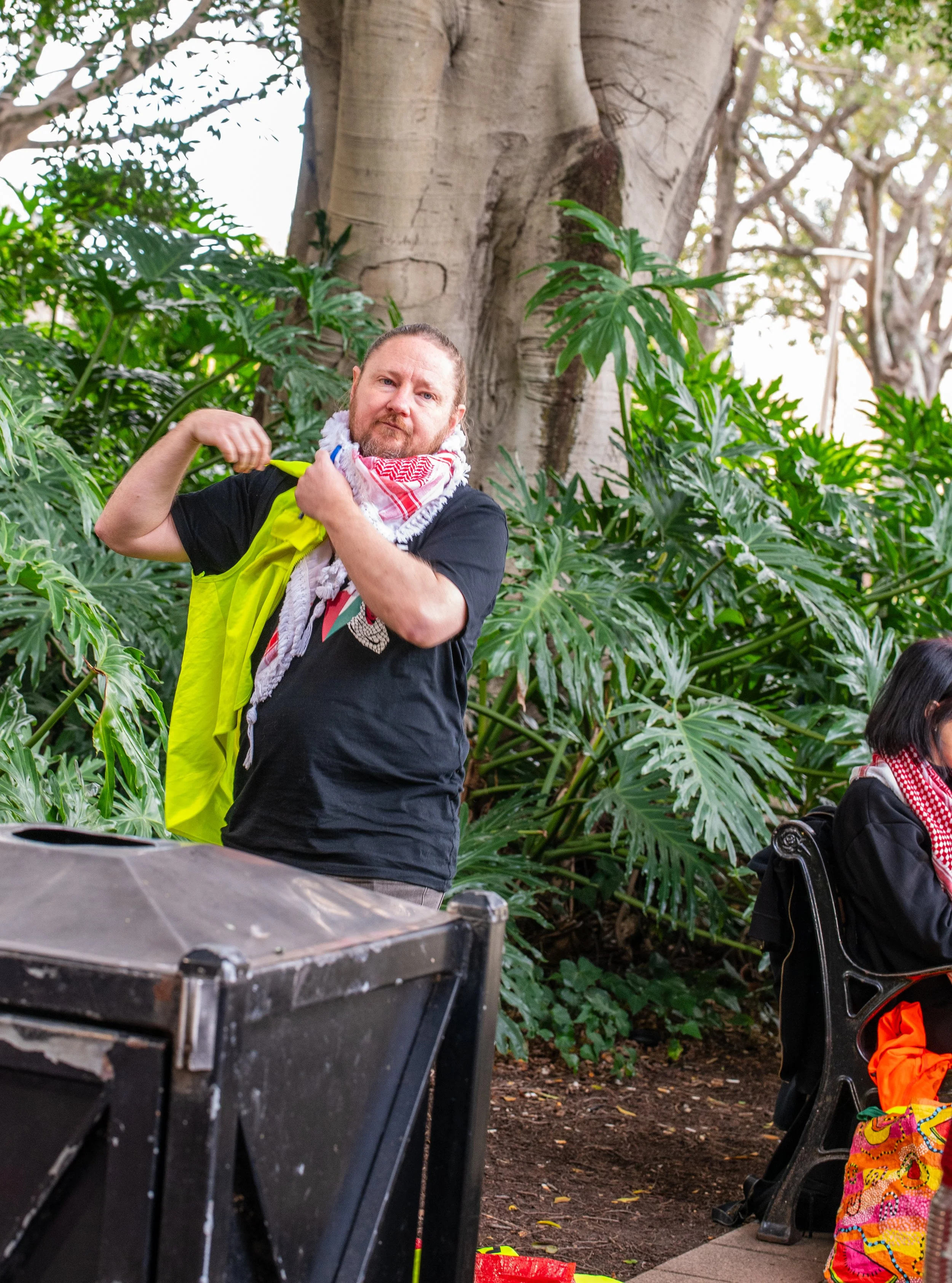 A man with a beard, wearing a black t-shirt, a red and white keffiyeh scarf, and a yellow vest, is standing outdoors among large green leafy plants and a big tree. He has his right arm raised and is pulling on the strap of his vest, looking at the camera.