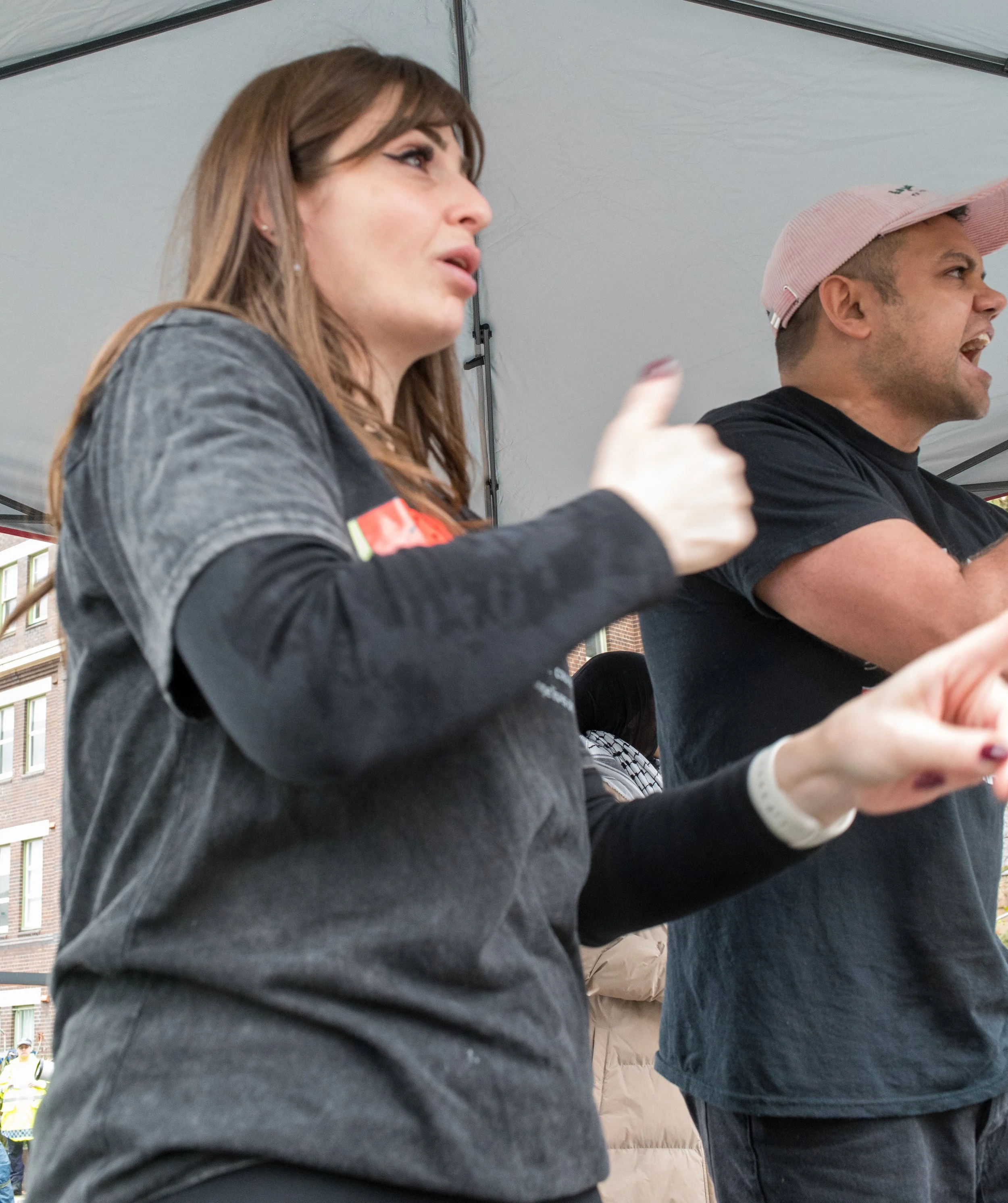 Two people standing at a booth under a canopy, talking passionately; the woman on the left has long brown hair, is wearing a dark gray shirt, and is giving a thumbs-up; the man on the right is wearing a pink cap and a black shirt.