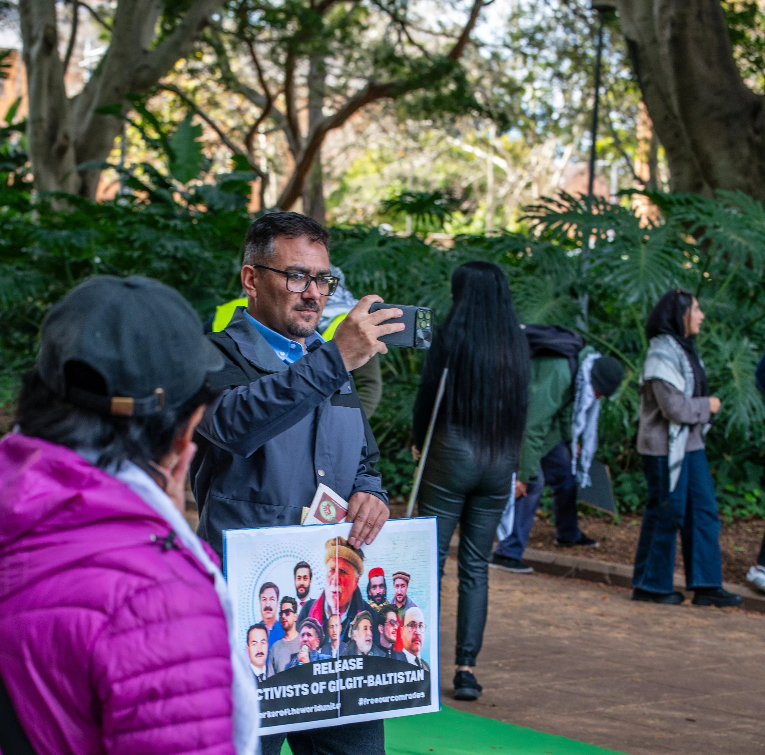 A man with glasses holding a smartphone and a sign protesting human rights violations in Gilgit-Baltistan, with other people standing in line outdoors among trees.