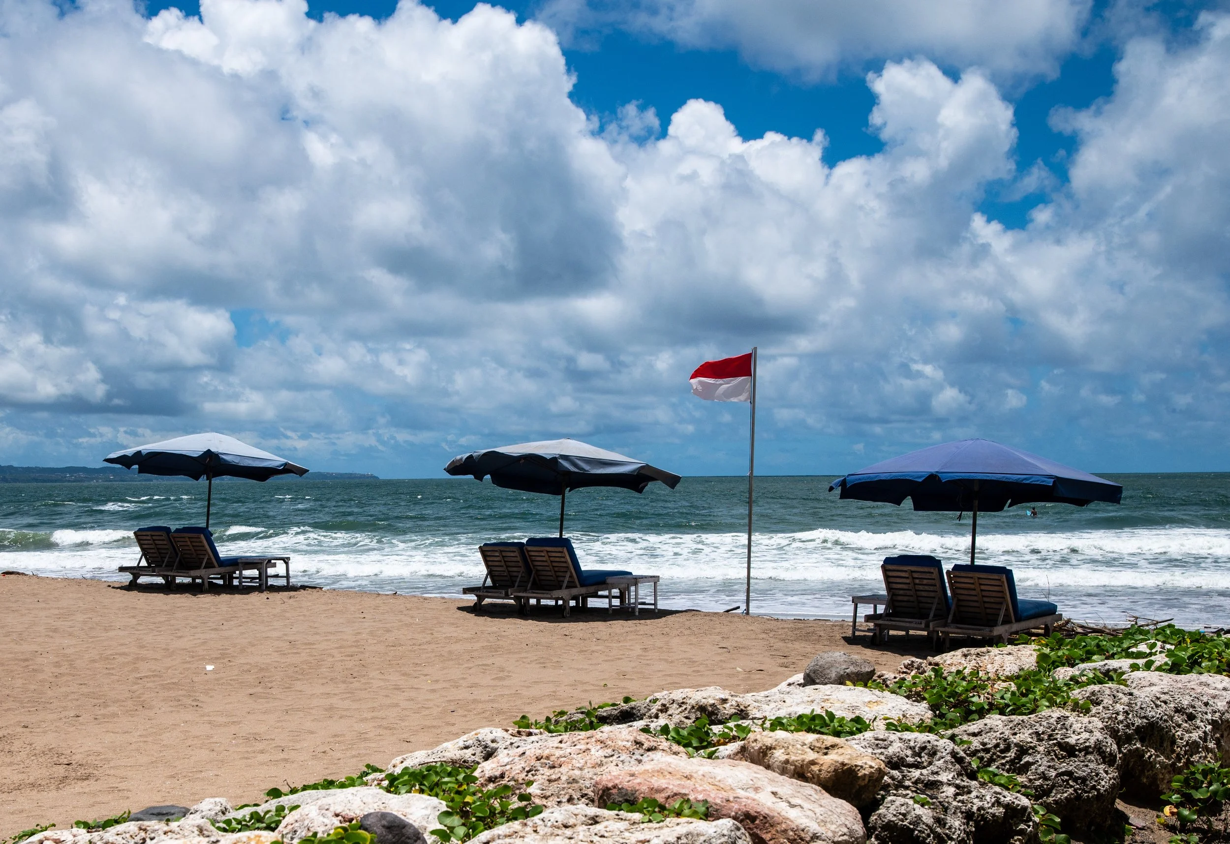 A beach with sand, blue and white umbrellas, blue chairs, a flag, and ocean waves under a partly cloudy sky.
