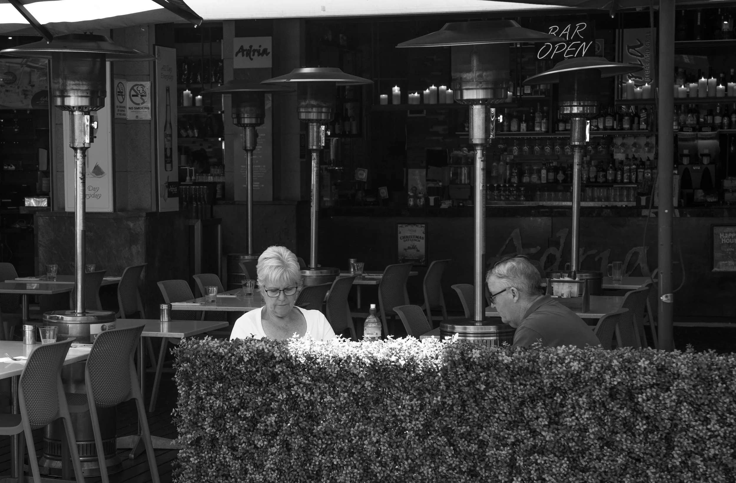 Two people sitting at a table outside a restaurant, separated by a hedge, with patio heaters overhead and a bar visible in the background.