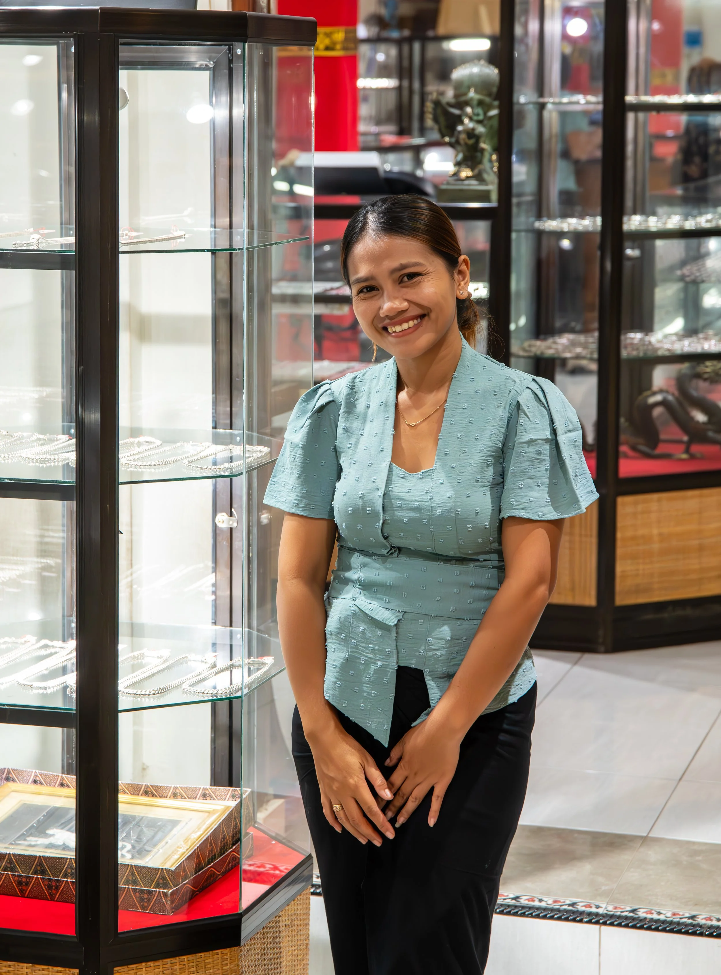 A smiling woman with dark hair, wearing a light teal blouse, stands next to glass display cases with jewelry inside in a store.