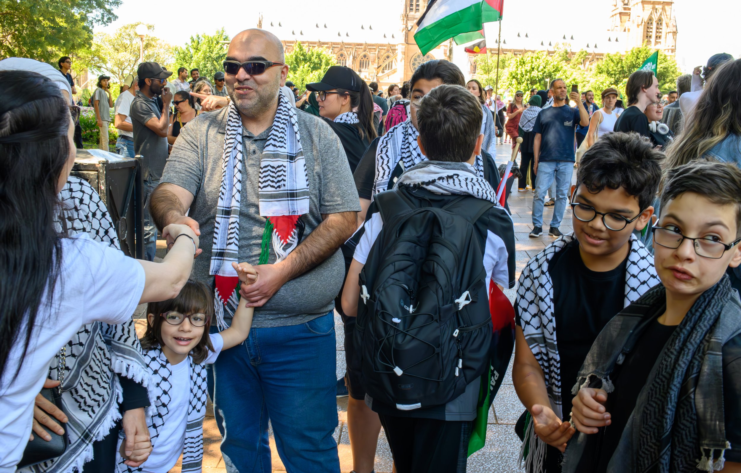 A group of people, including children and adults, are gathered outdoors at a protest or rally. The man in the center is shaking hands with a woman. Many individuals are wearing scarves with the Palestinian flag pattern. In the background, there is a building that appears to be a church or cathedral, and others are holding flags, including the Palestinian flag.