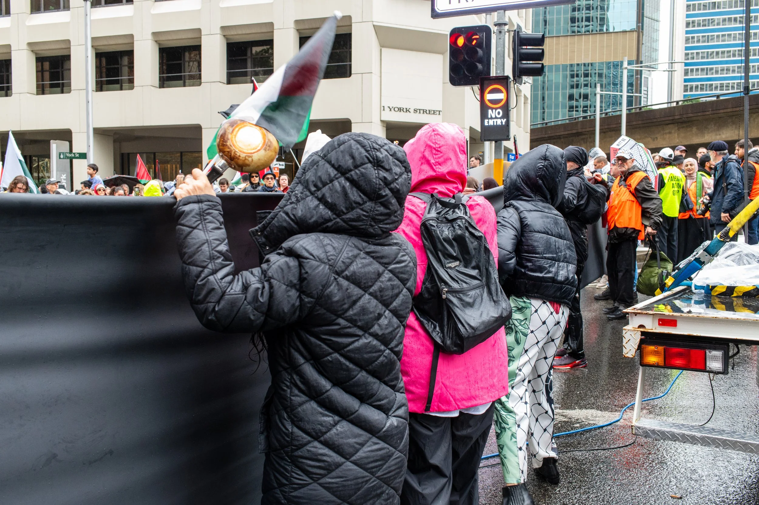 People standing in line outdoors during a protest or demonstration, some holding flags and banners, with a city street and buildings in the background on a rainy day.