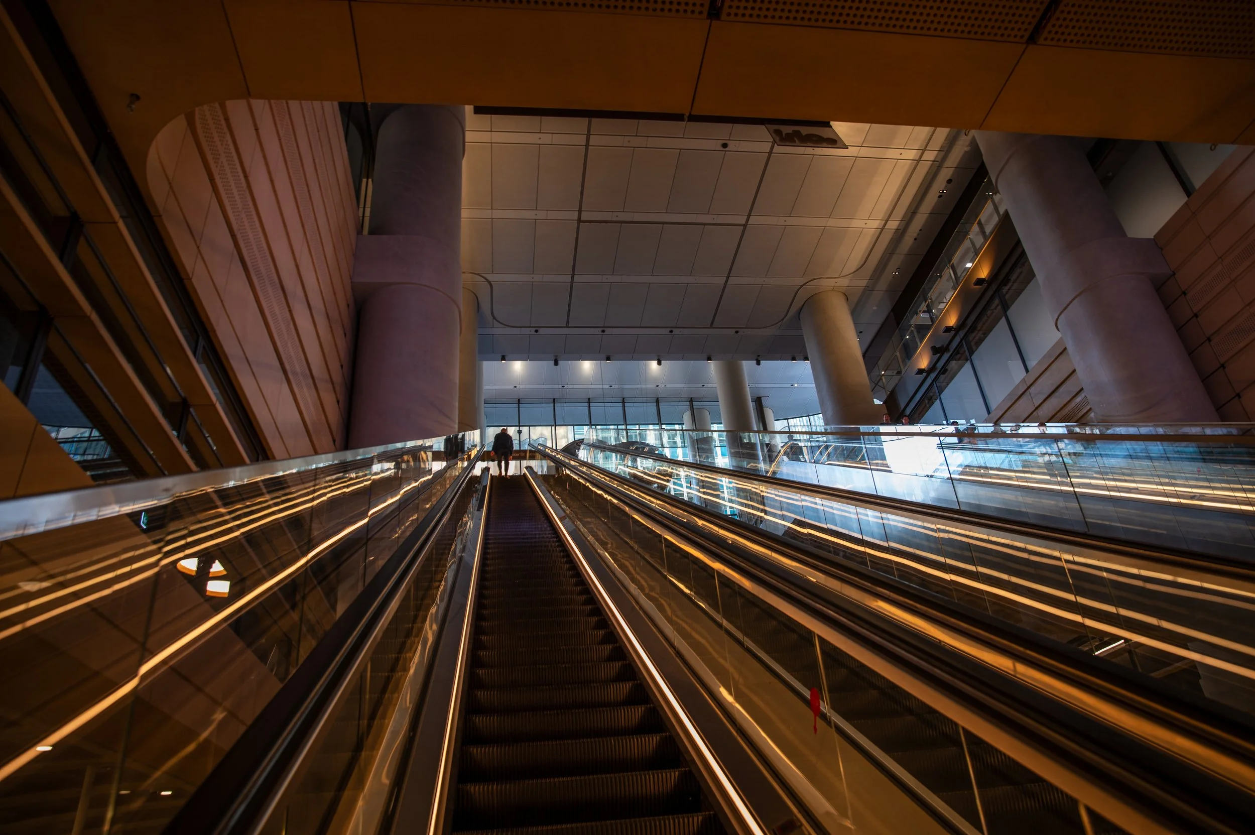 Interior of a modern building with an escalator leading upward, large concrete columns, and glass walls allowing natural light to fill the space.