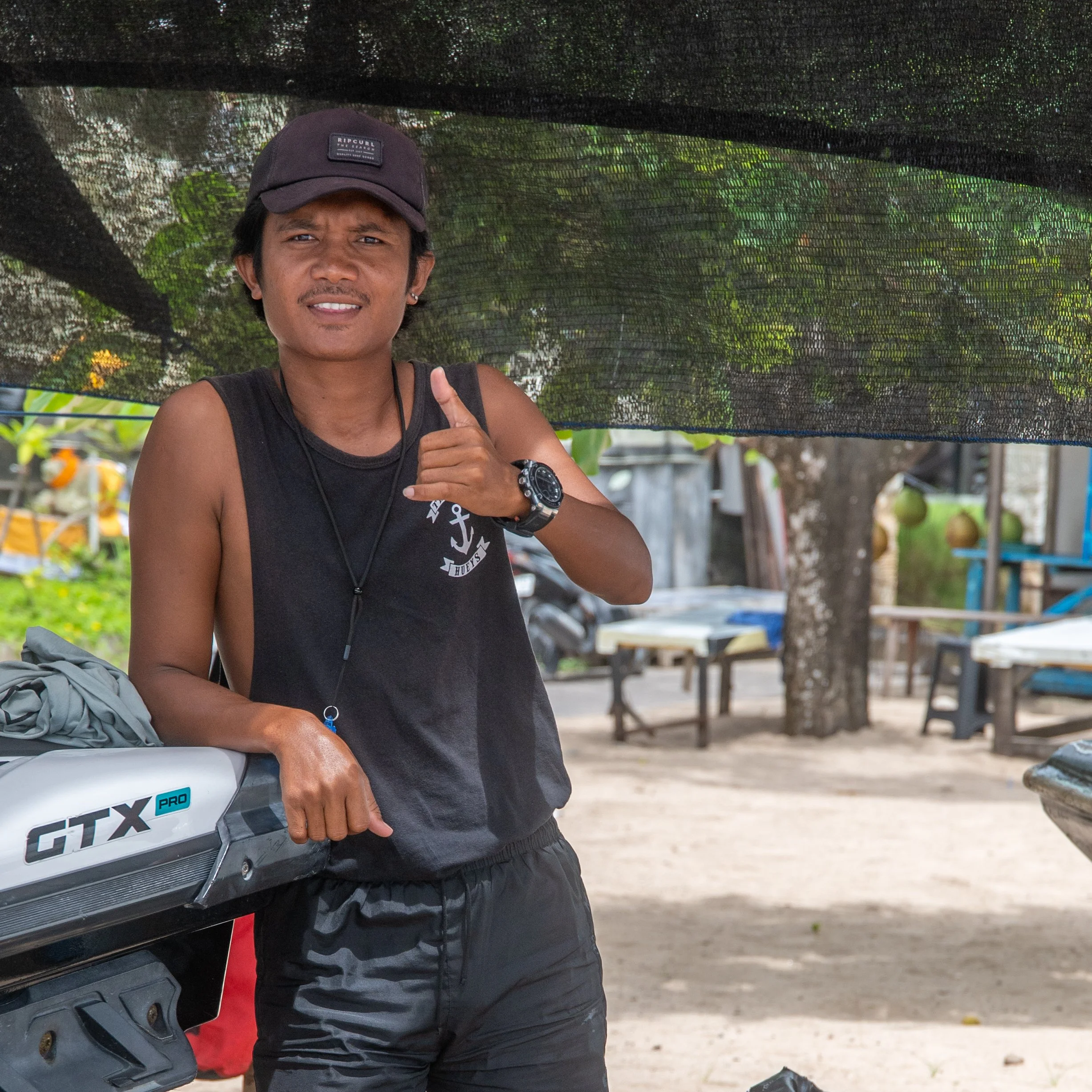 A young man wearing a black cap, black sleeveless shirt, and black watch giving a thumbs-up while standing outside under a shade cloth, holding a piece of equipment or a box with the label 'GTX PRO'.