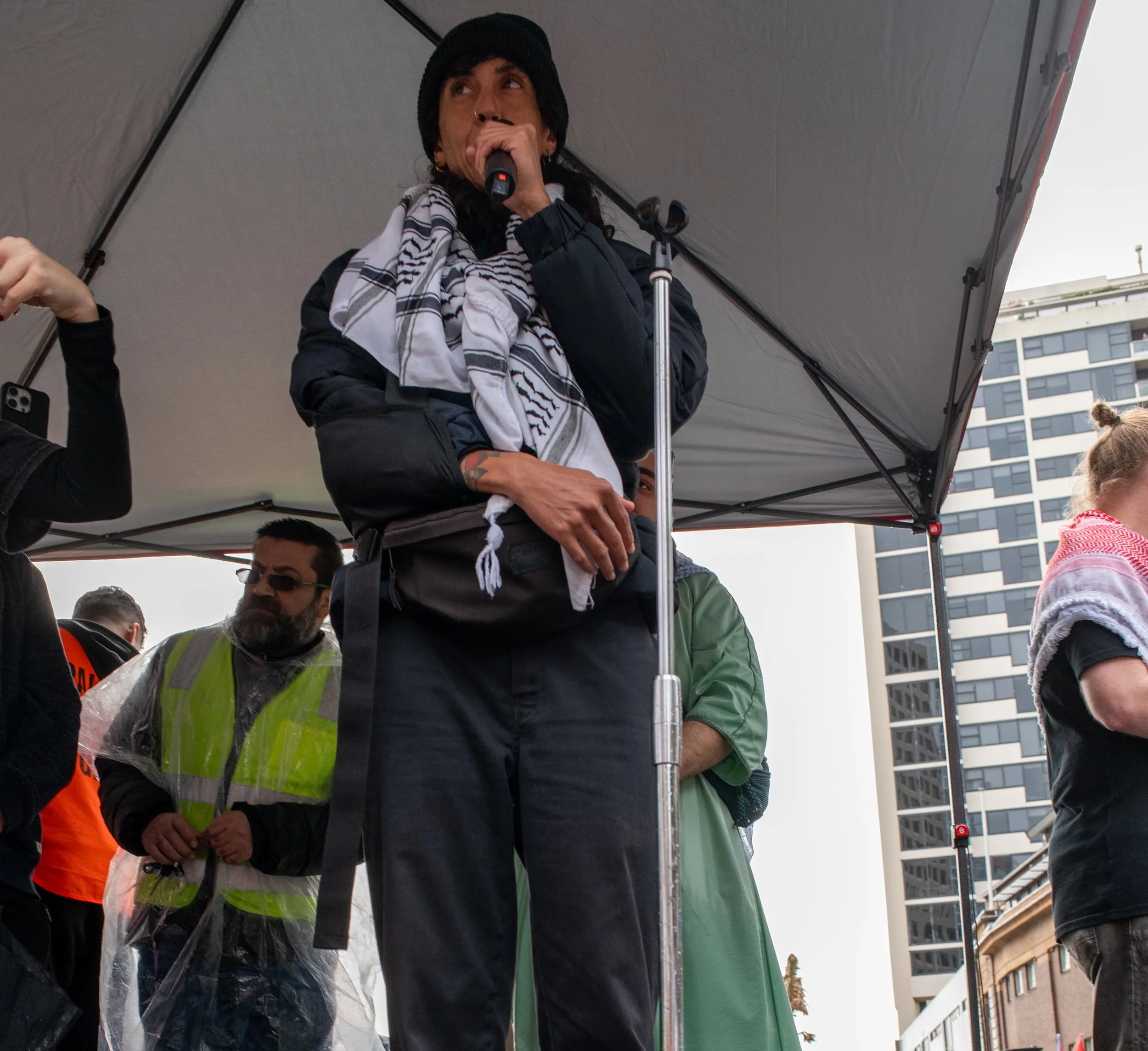 A woman speaking into a microphone at a public gathering, wearing a black beanie, a black jacket, and a white scarf with black stripes. She is under a canopy, with city buildings in the background.