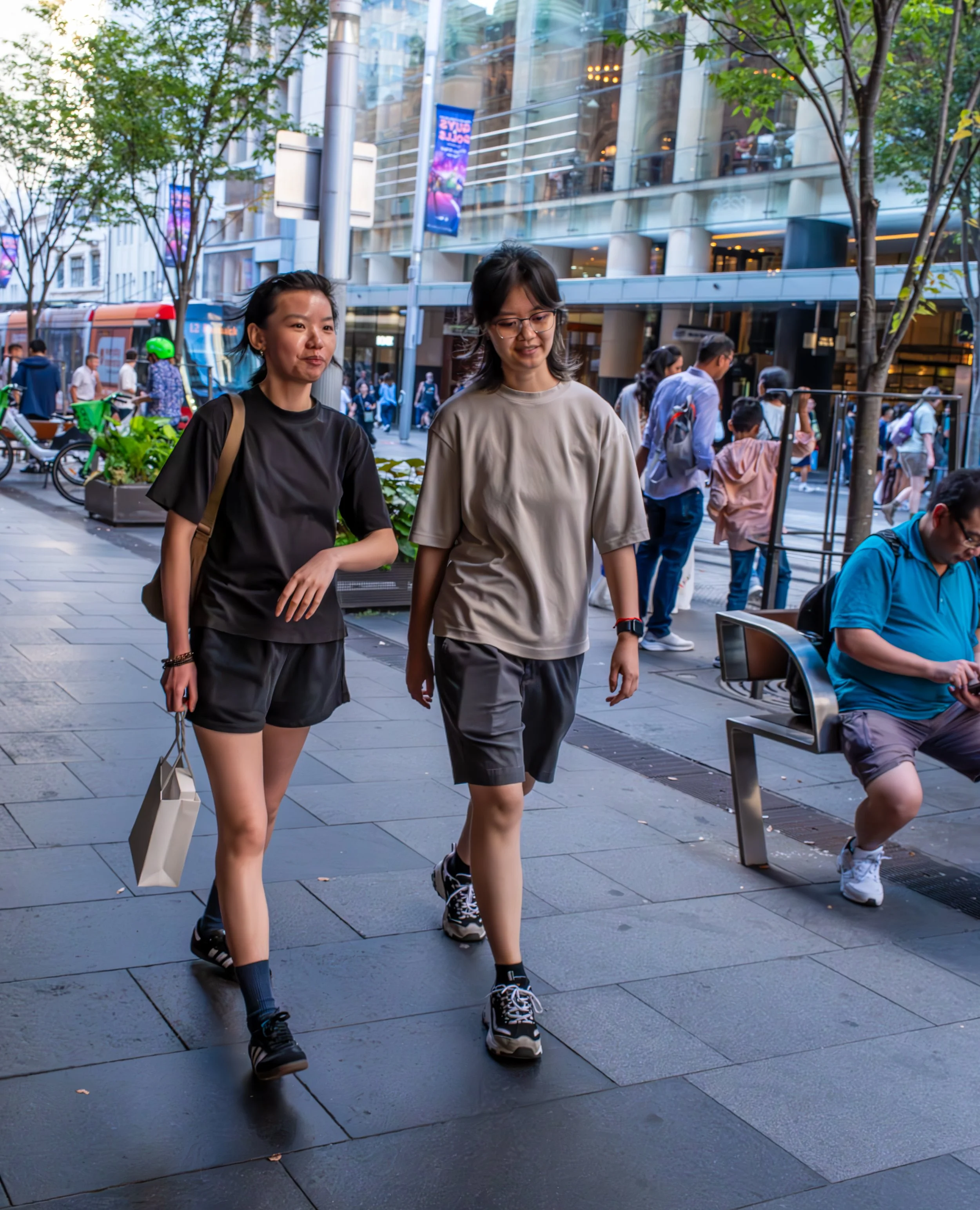 Two young women walking on a city sidewalk amidst pedestrians with modern buildings and trees in the background.