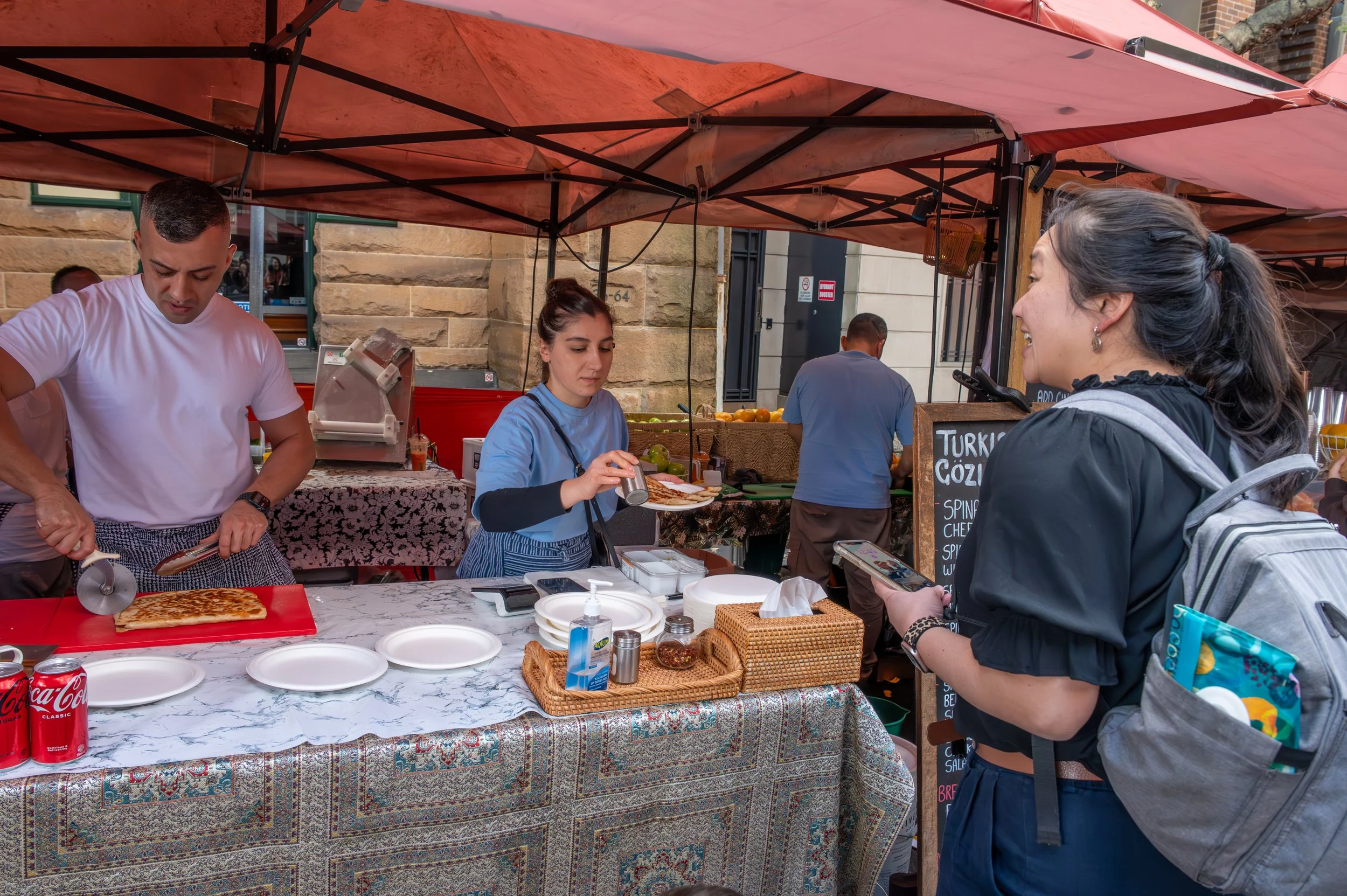 A woman is ordering food at a street food stall, where a man is preparing a flatbread. The woman is smiling while holding a phone, and another woman behind the counter is adding condiments. The stall is set up under a red canopy with various kitchen items and a menu board nearby.