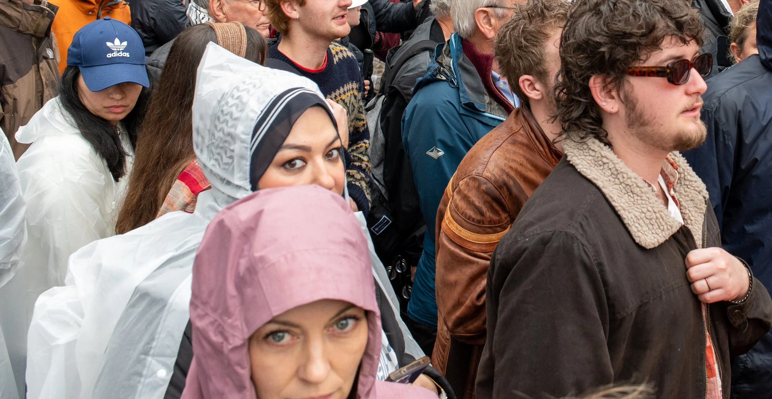 Group of diverse people standing outdoors, some wearing rain gear and jackets, with focused or neutral expressions.