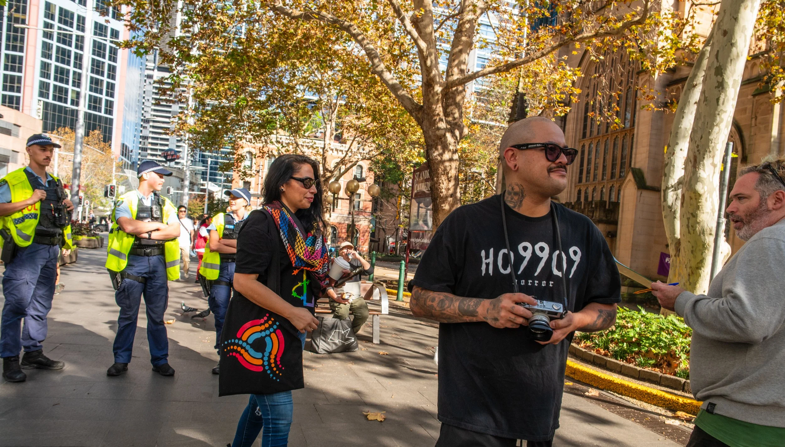 Group of police officers and civilians engaged in conversation outdoors in an urban park with trees and tall buildings in the background.