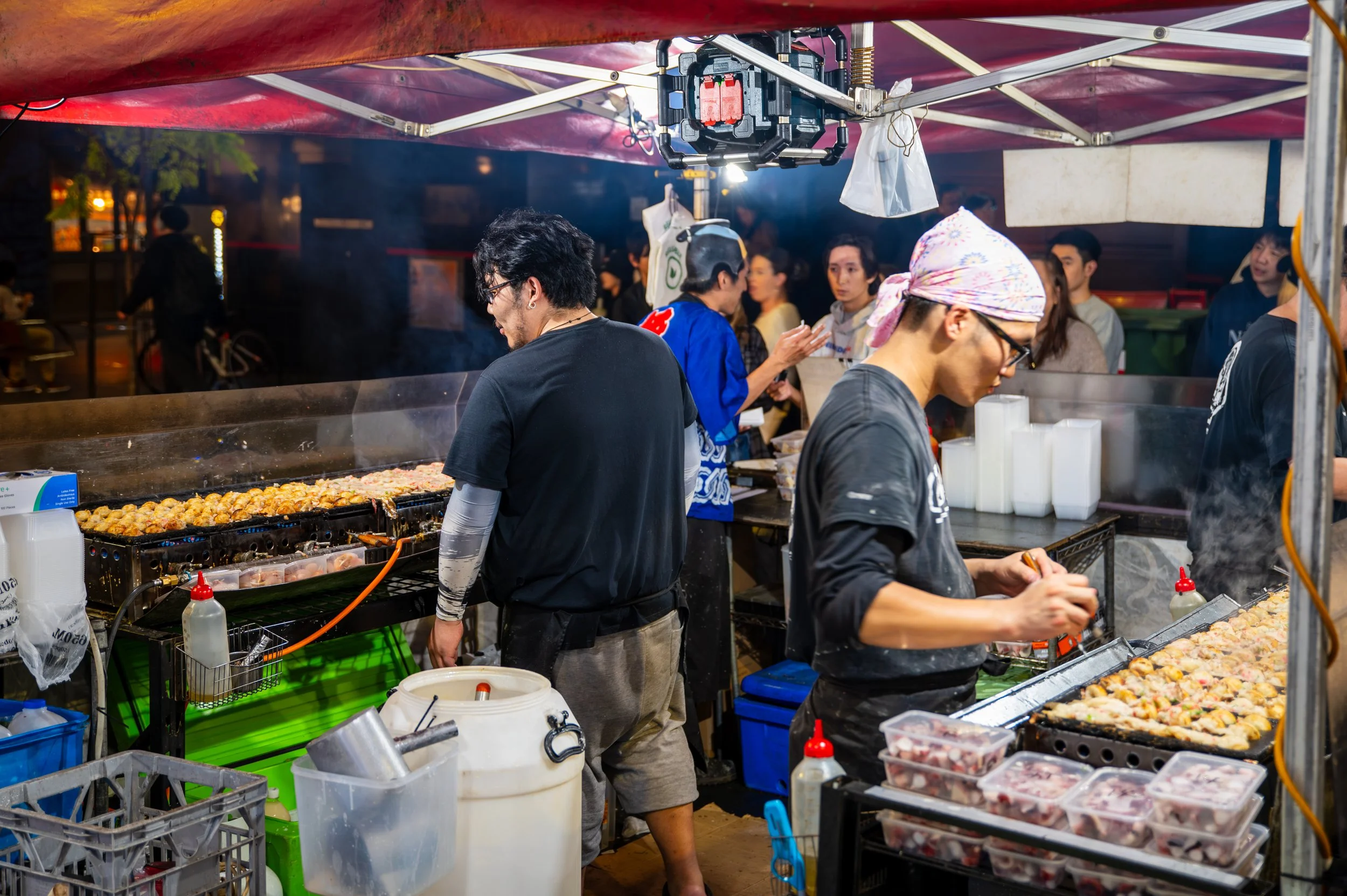 Two food vendors preparing takoyaki at a night market stall, with a crowd of customers in the background.