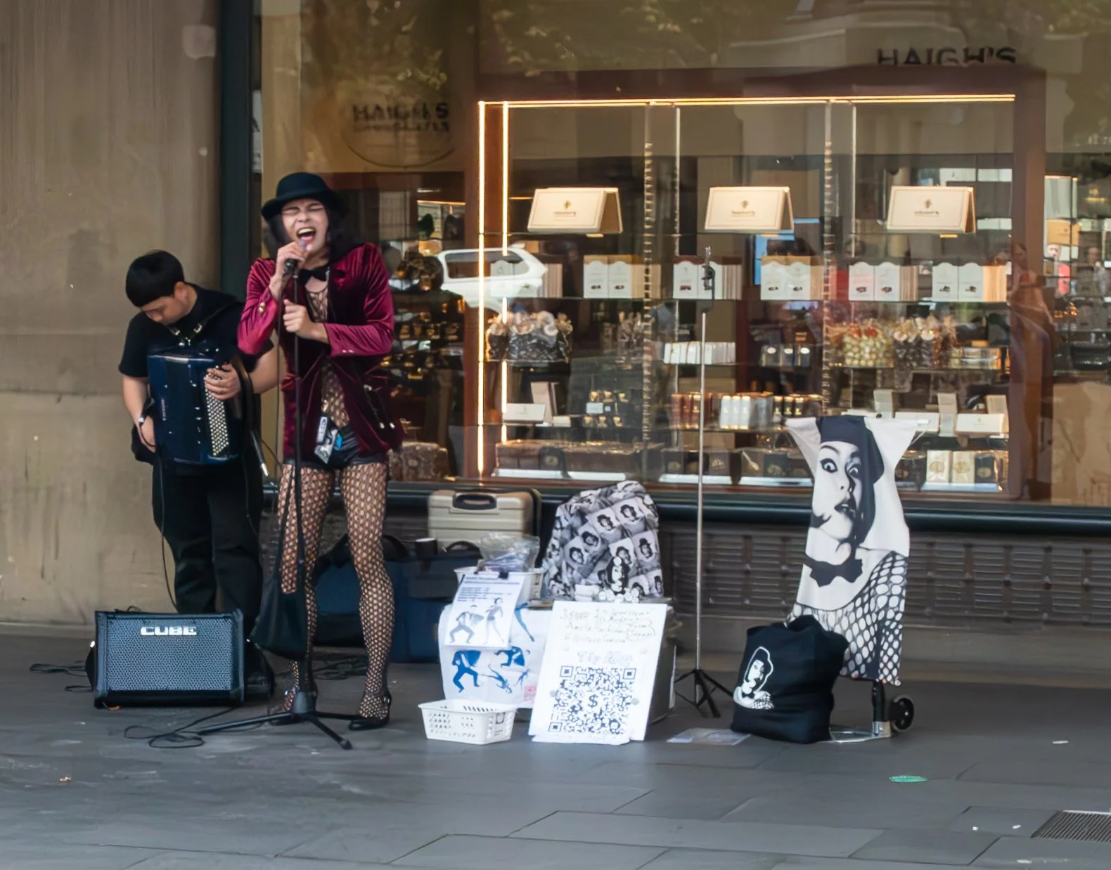 Street performer singing into a microphone with a boy playing an accordion beside her, set up in front of a store window displaying chocolates and gifts, with QR code and artwork on display.