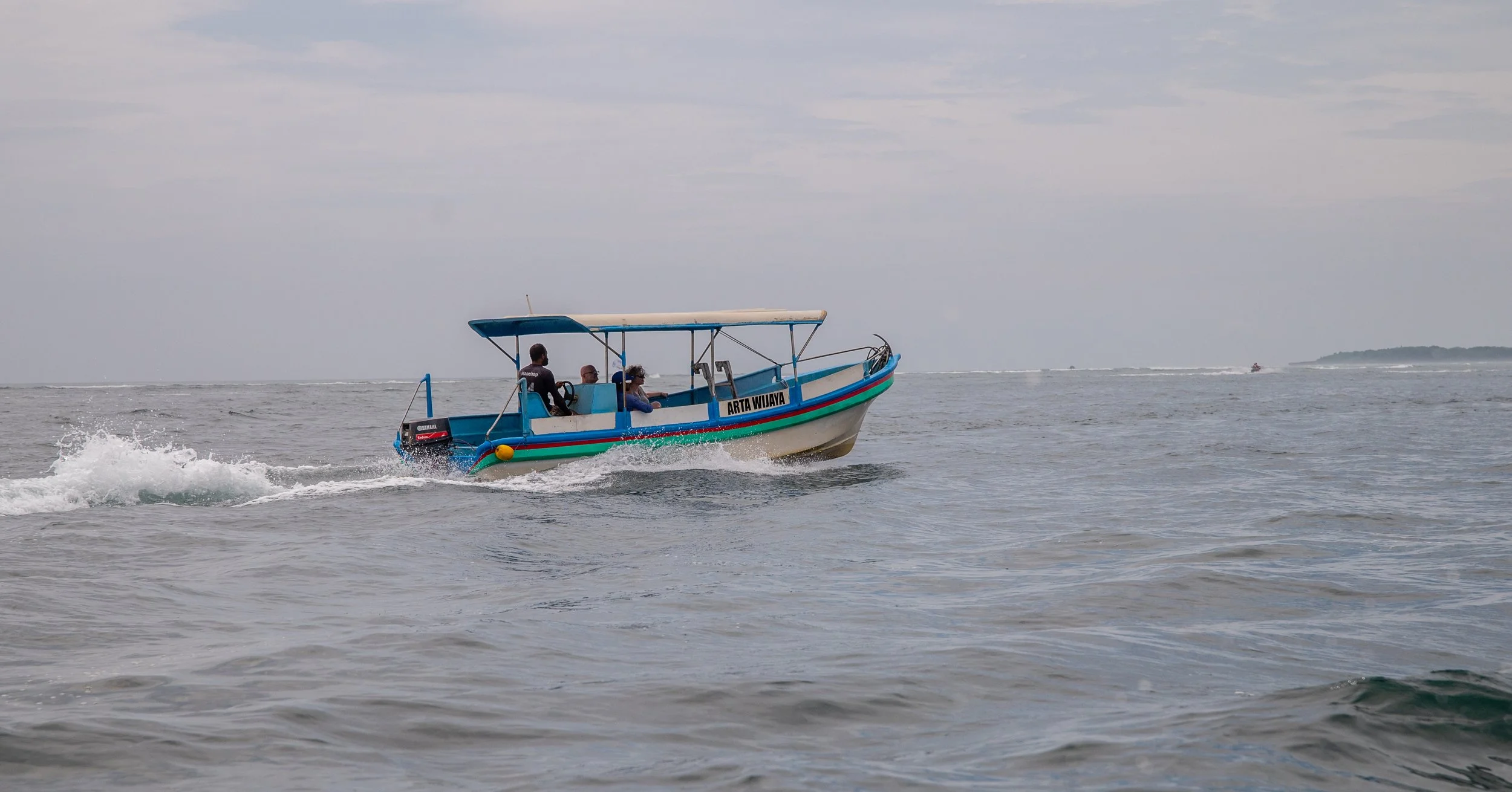 A small blue and white motorboat with four people aboard speeding across the water, leaving a wake behind. The boat has a canopy and the name 'ARTA WIJAYA' on the side.
