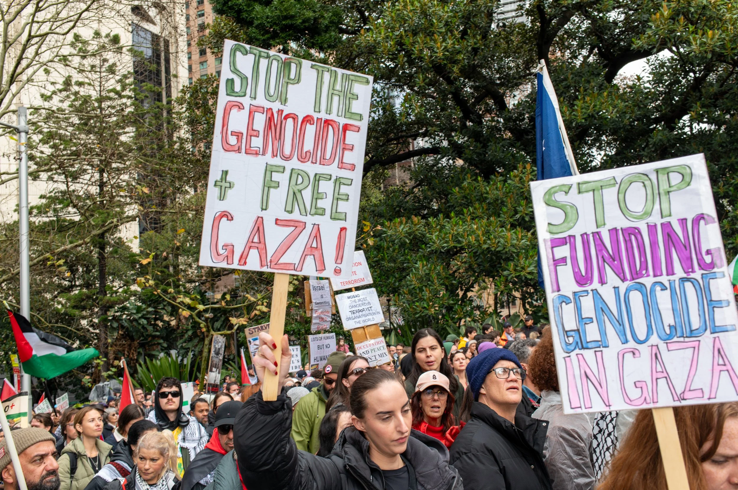 Crowd of protesters holding signs at a rally, with trees and buildings in the background. Signs include messages like "Stop the genocide + free Gaza!" and "Stop funding genocide in Gaza".