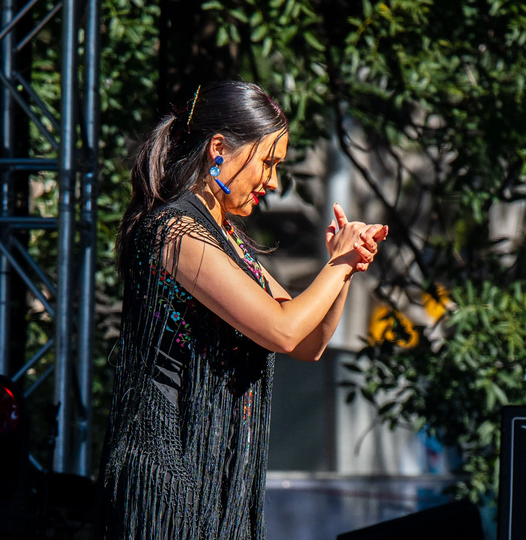 A woman with dark hair styled in a ponytail, wearing colorful beaded jewelry and a black fringe outfit, is performing a dance or gesture on stage outdoors.