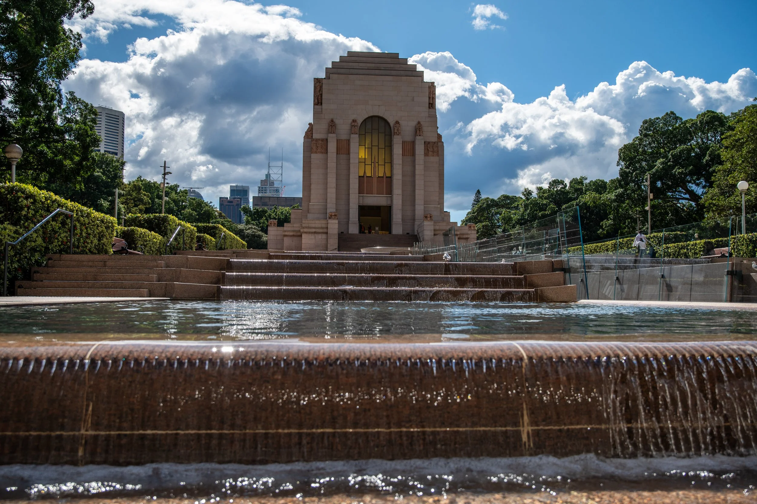 A memorial with a large central building, cascading water feature in the foreground, greenery on both sides, and a partly cloudy sky above.