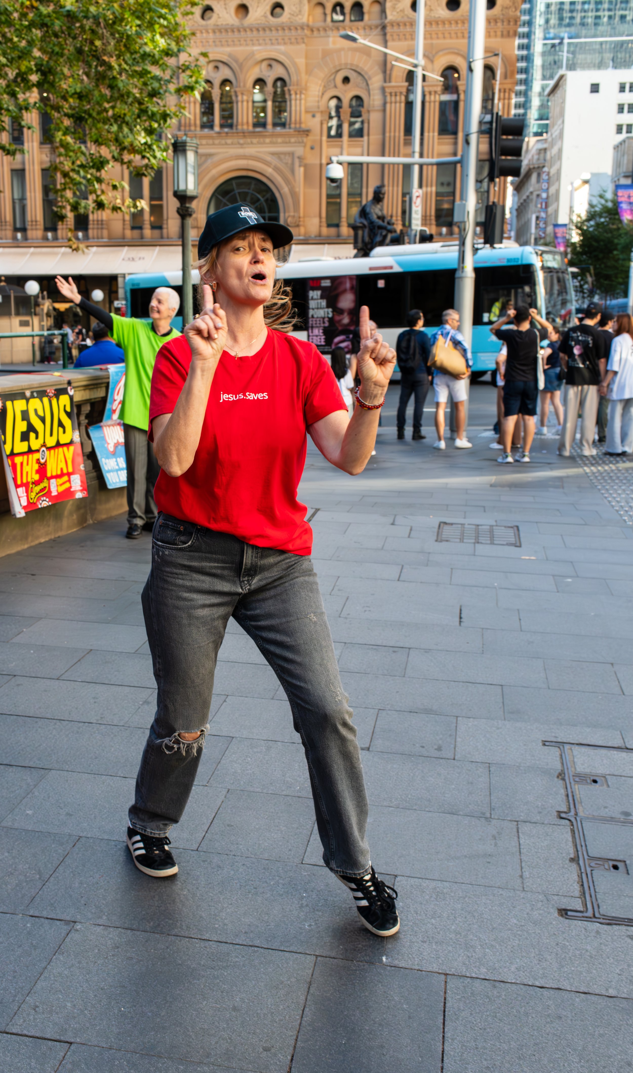 A woman in a red t-shirt and baseball cap is speaking passionately and gesturing with her hands at a busy city intersection, with people and a bus in the background.