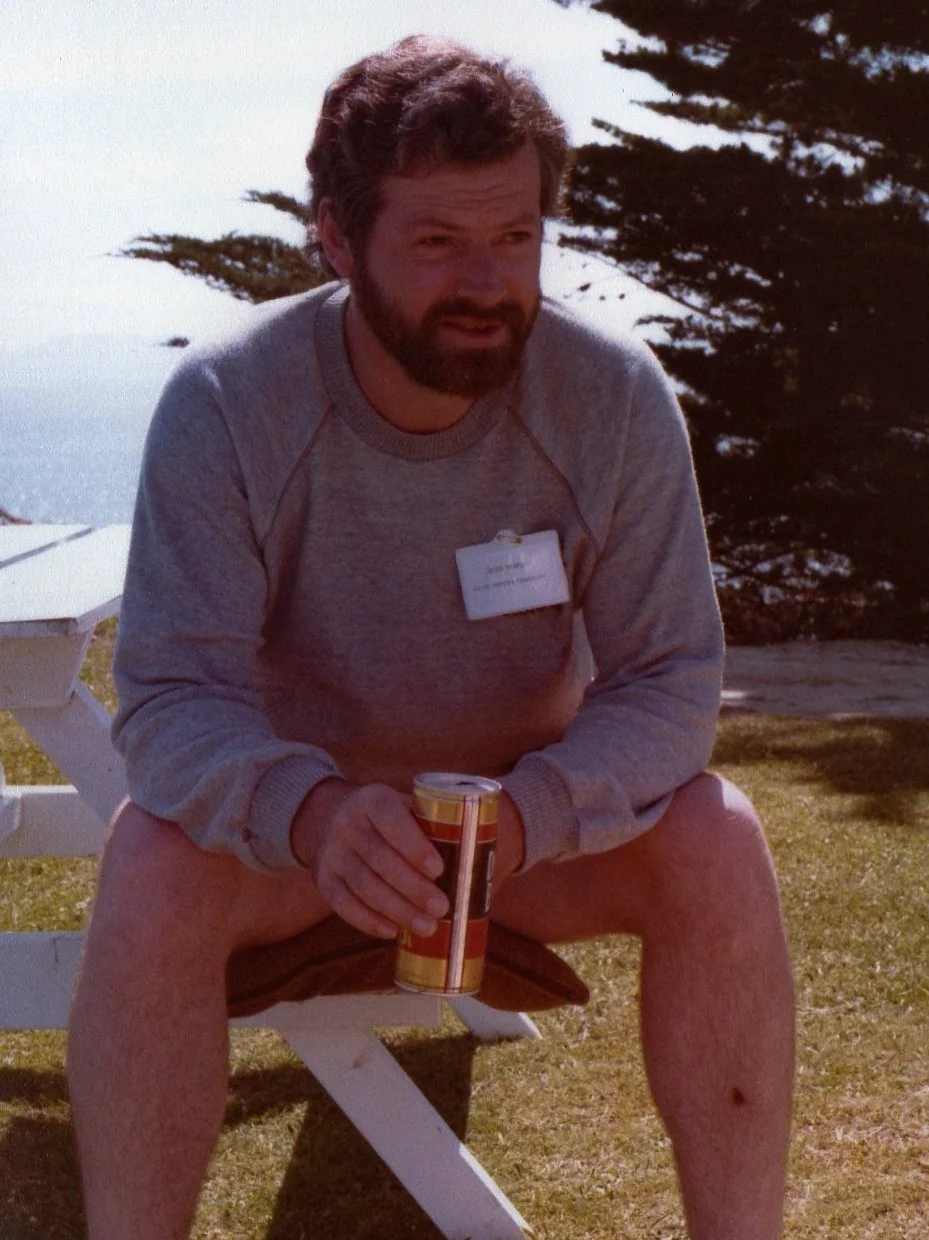 A man with a beard and curly hair, sitting outdoors on a white bench near the water, holding a can of soda, wearing a gray sweatshirt and shorts, with a park badge on his chest, in a nature setting with trees and water in the background.