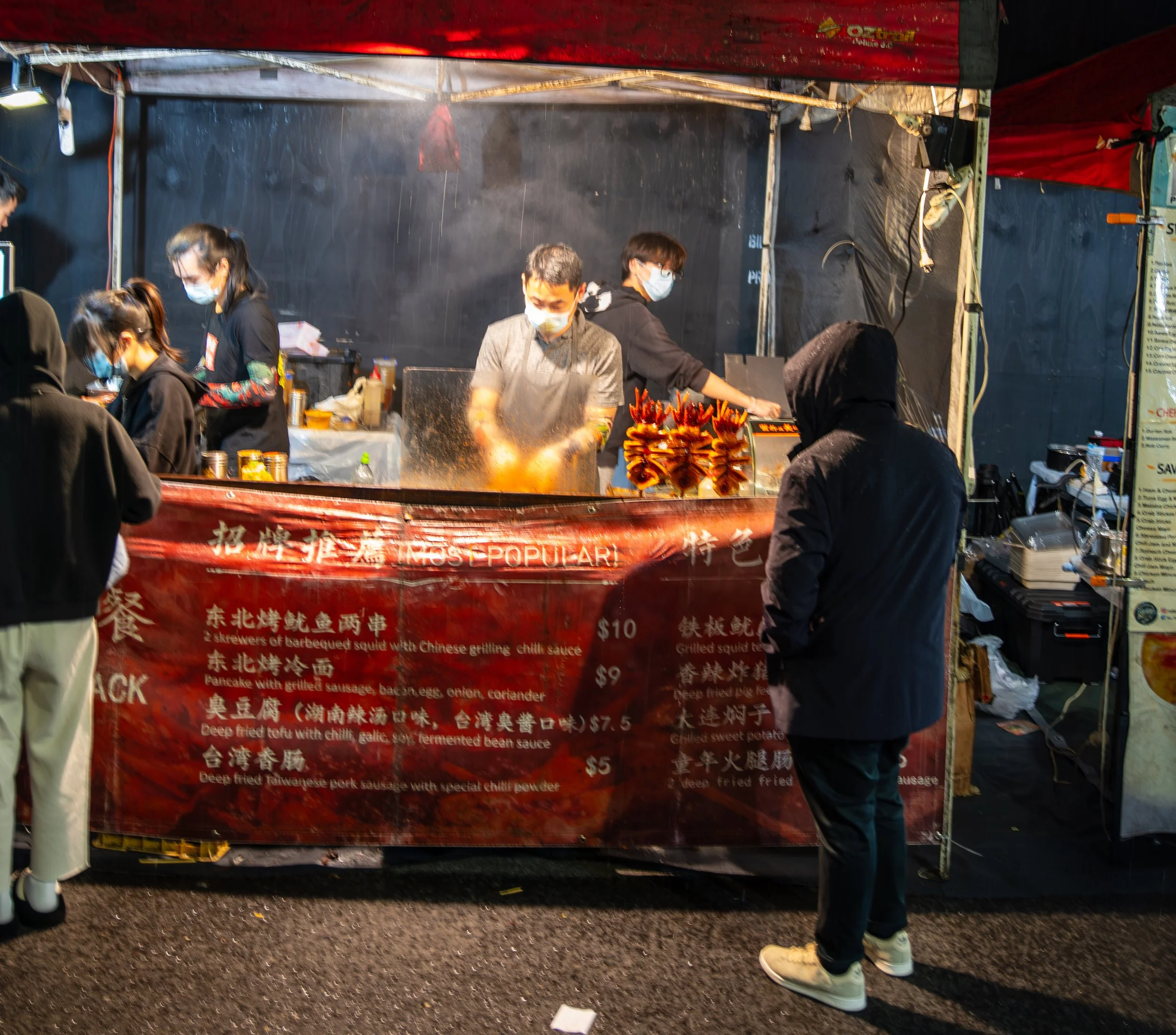 Food stall at a night market with vendors cooking and customers ordering. The stall offers skewers of barbequed squid, pancakes with sausage, and deep-fried tofu, with prices listed on a red sign. Customers and vendors are wearing masks.
