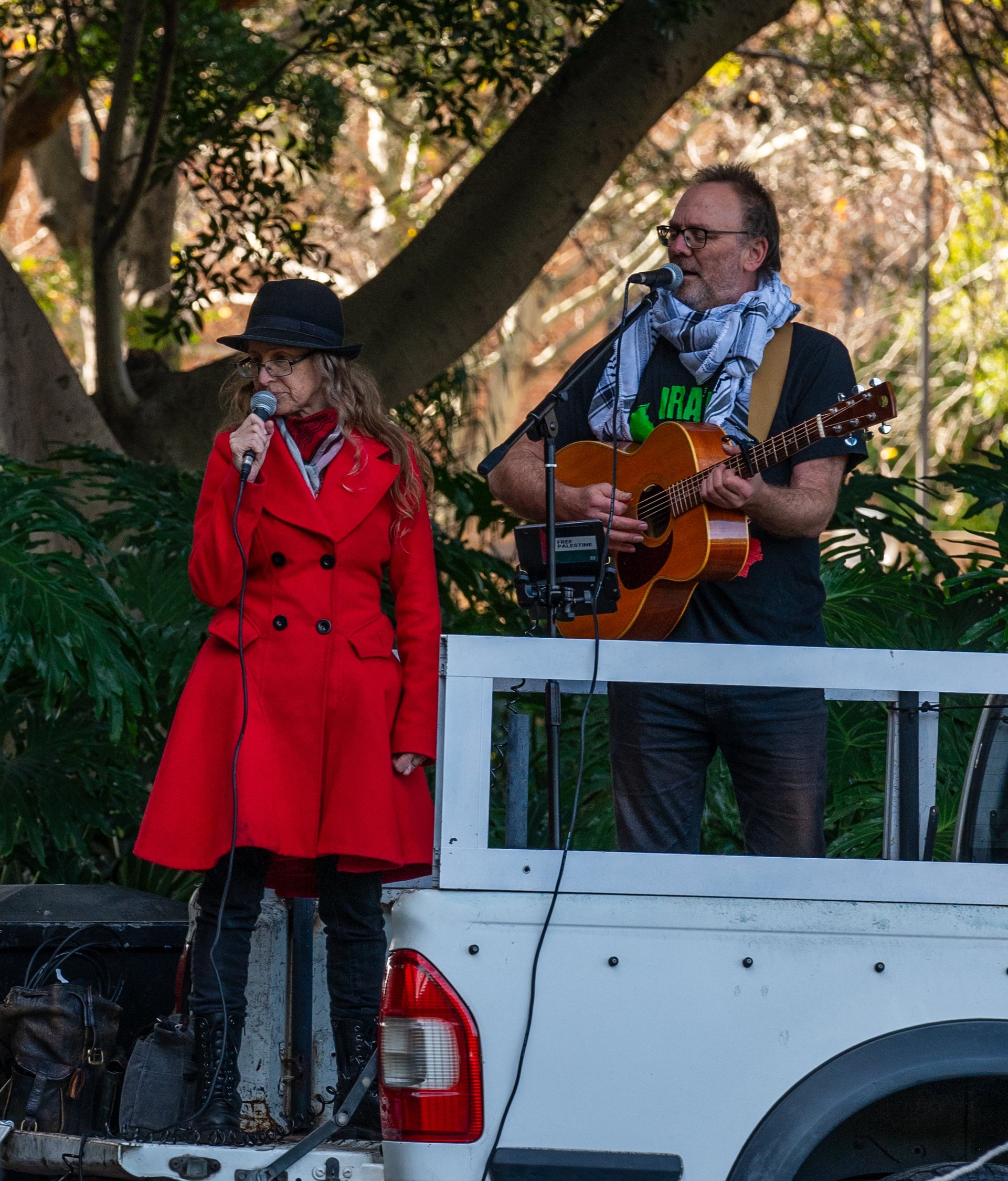 A man and a woman performing music on a small stage outdoors, with the man playing an acoustic guitar and the woman singing into a microphone. The woman is wearing a red coat and black hat, and the man is wearing glasses, a scarf, and a black t-shirt. The stage is set on the back of a white pickup truck with a background of trees and greenery.