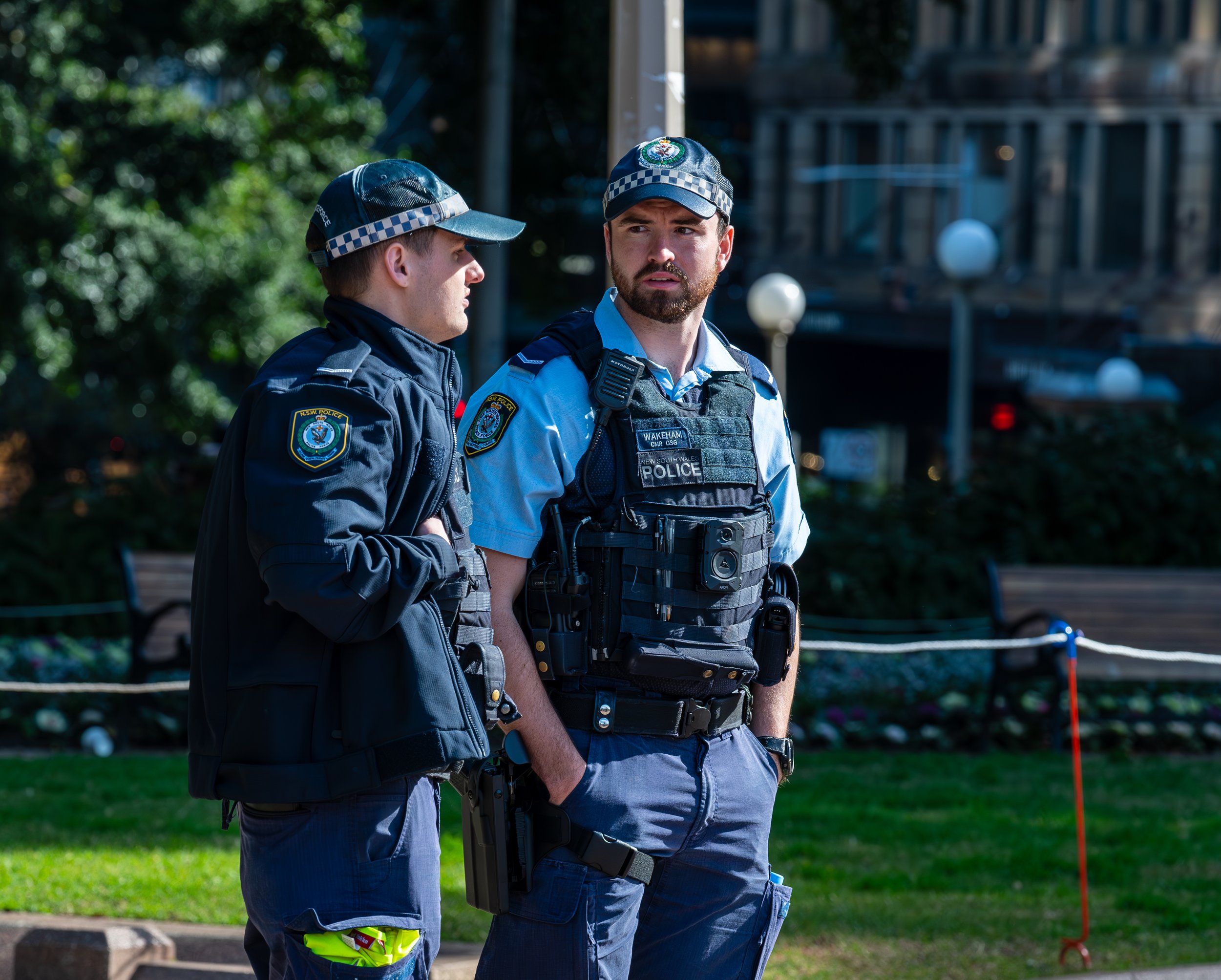 Two police officers stand conversing outdoors in a park-like setting, both wearing uniforms and tactical gear.