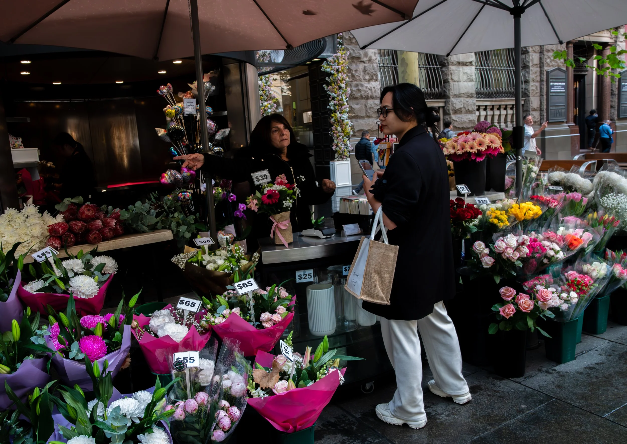 A woman in white pants and a black coat shopping for flowers at a busy outdoor flower stand, with various colorful flowers and price tags displayed.