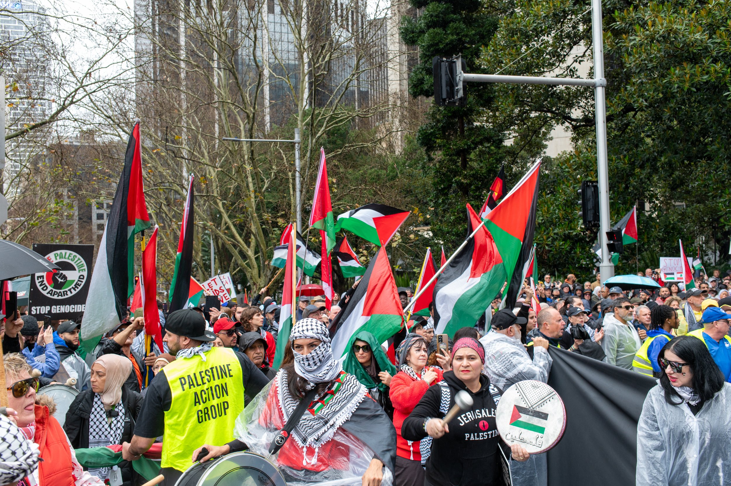 Crowd of protesters holding Palestinian flags and signs, wearing Palestinian scarves and clothing, participating in a demonstration outdoors with trees and buildings in the background.
