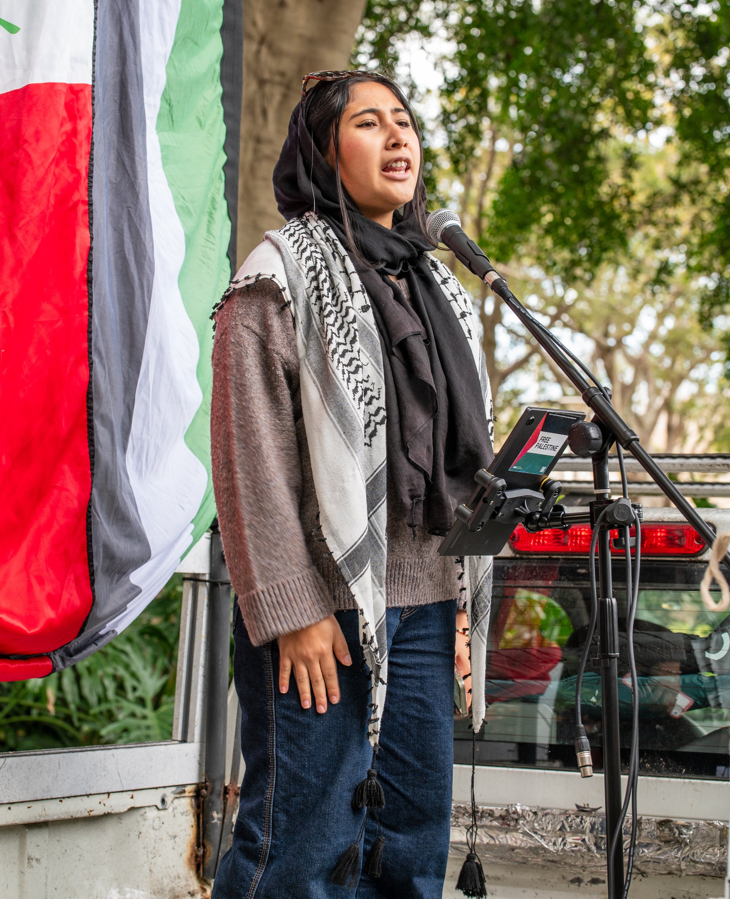 A woman speaking at a rally or protest, with a Palestinian flag behind her, outdoors with trees in the background.