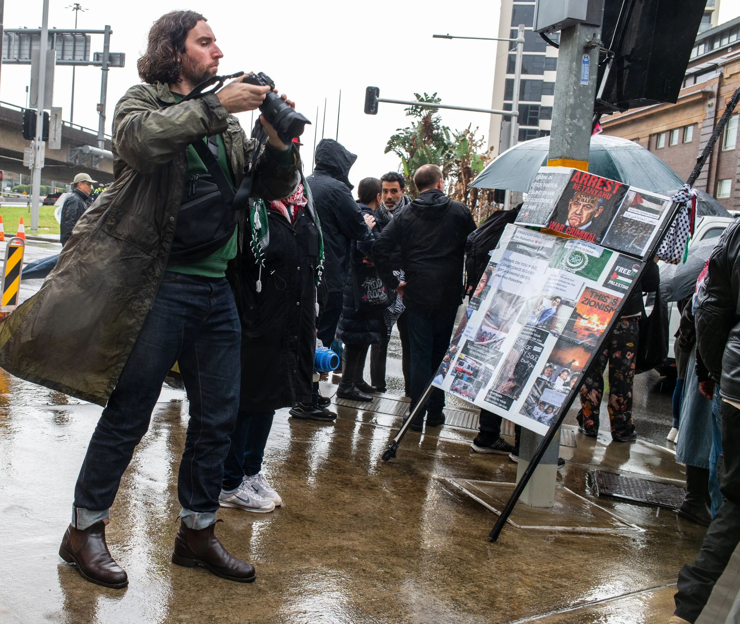 A man with long hair taking a photo with a camera on a rainy day at a protest or rally, with a crowd of people and protest signs displayed on a board.