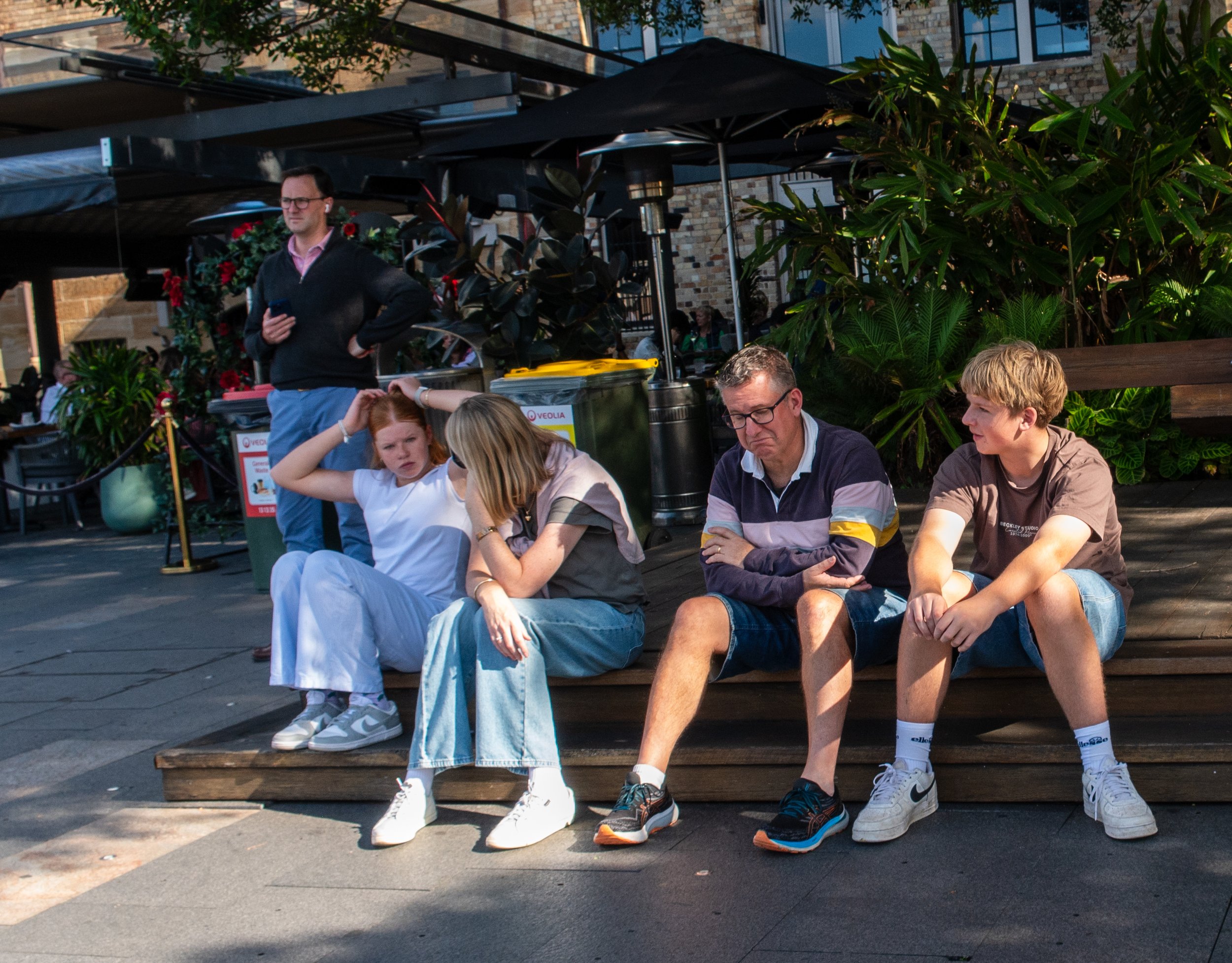 Four people sitting on a wooden ledge outdoors, two women and two young men, with a man standing behind them. One woman has red hair and is touching her head, and the other has blonde hair and is leaning in towards her. The young men have blonde and brown hair, sitting with crossed arms. Behind them, there are green plants, a building, and a person looking at their phone.