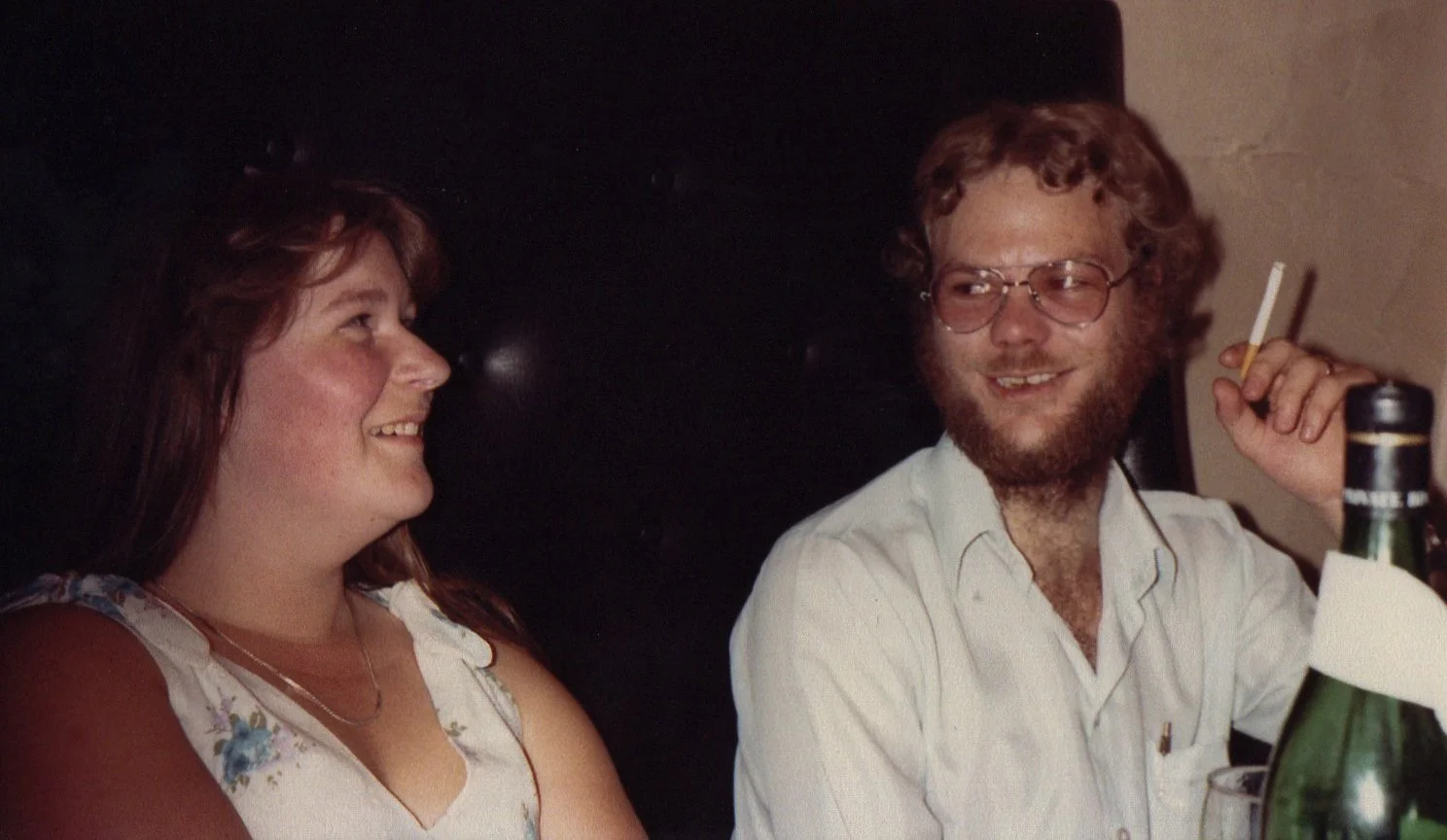 A young woman and man sitting at a table, smiling and talking. The man is holding a cigarette and has a beer bottle in front of him.